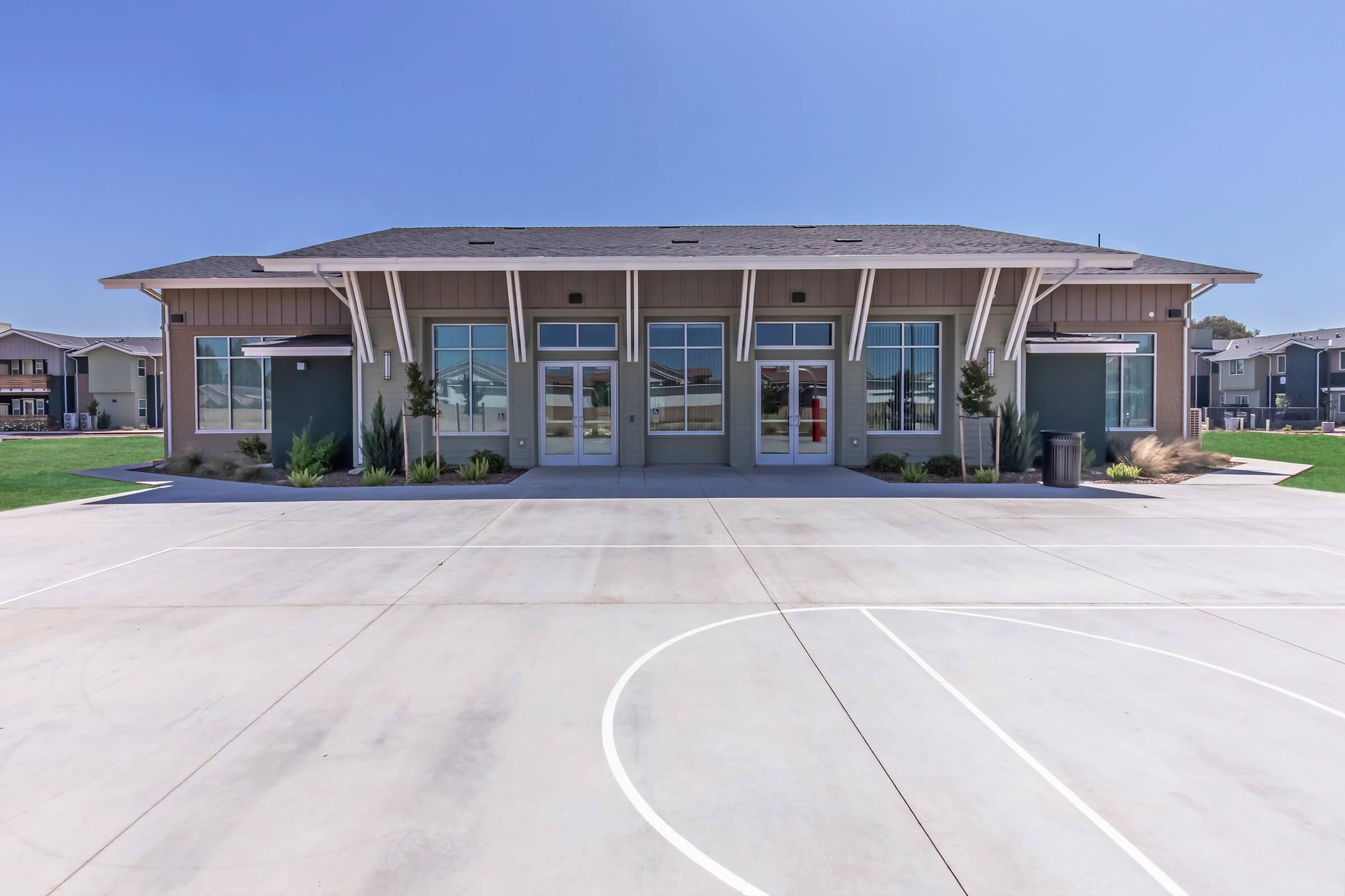 A modern community center featuring a wide entrance with large glass doors, surrounded by well-maintained landscaping. The building has a light-colored exterior and is set against a clear blue sky. In front, there is a basketball court with a smooth concrete surface.
