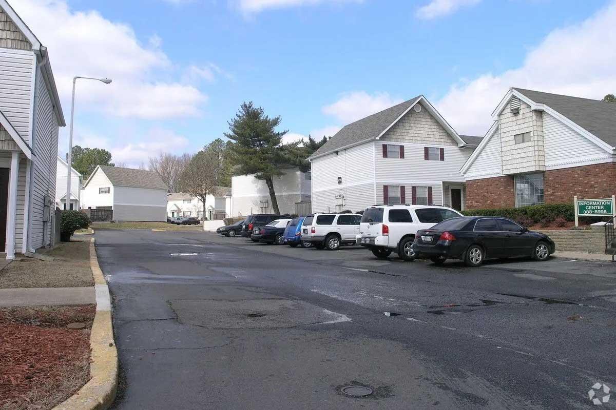 A residential street with several white and brick houses on either side, parked cars lining the street, and a clear blue sky with clouds. A sign for an apartment complex is visible, along with green landscaping and a tree in the background. The road is paved and has a slight curve.