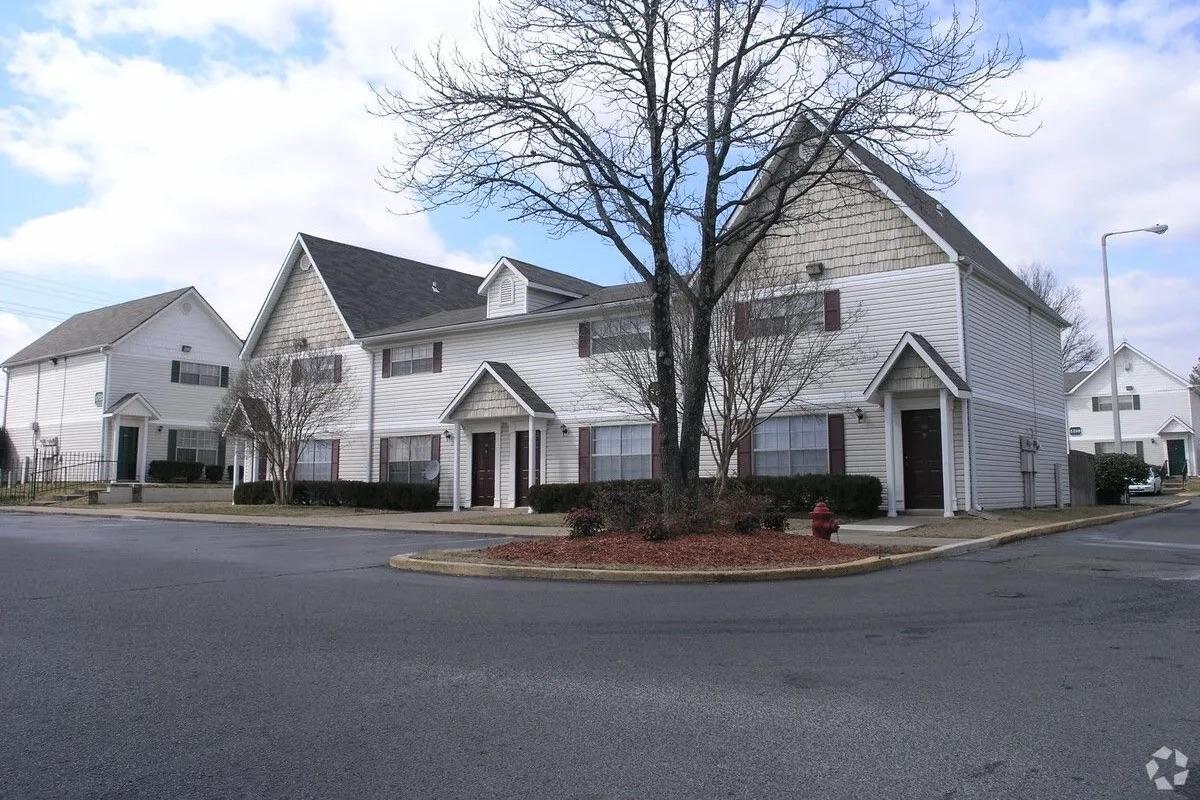 A corner view of a residential building featuring two stories, with a mix of gabled roofs and siding. The setting includes landscaped areas with shrubs and a fire hydrant. The sky is partly cloudy, and there are no visible cars or people nearby.