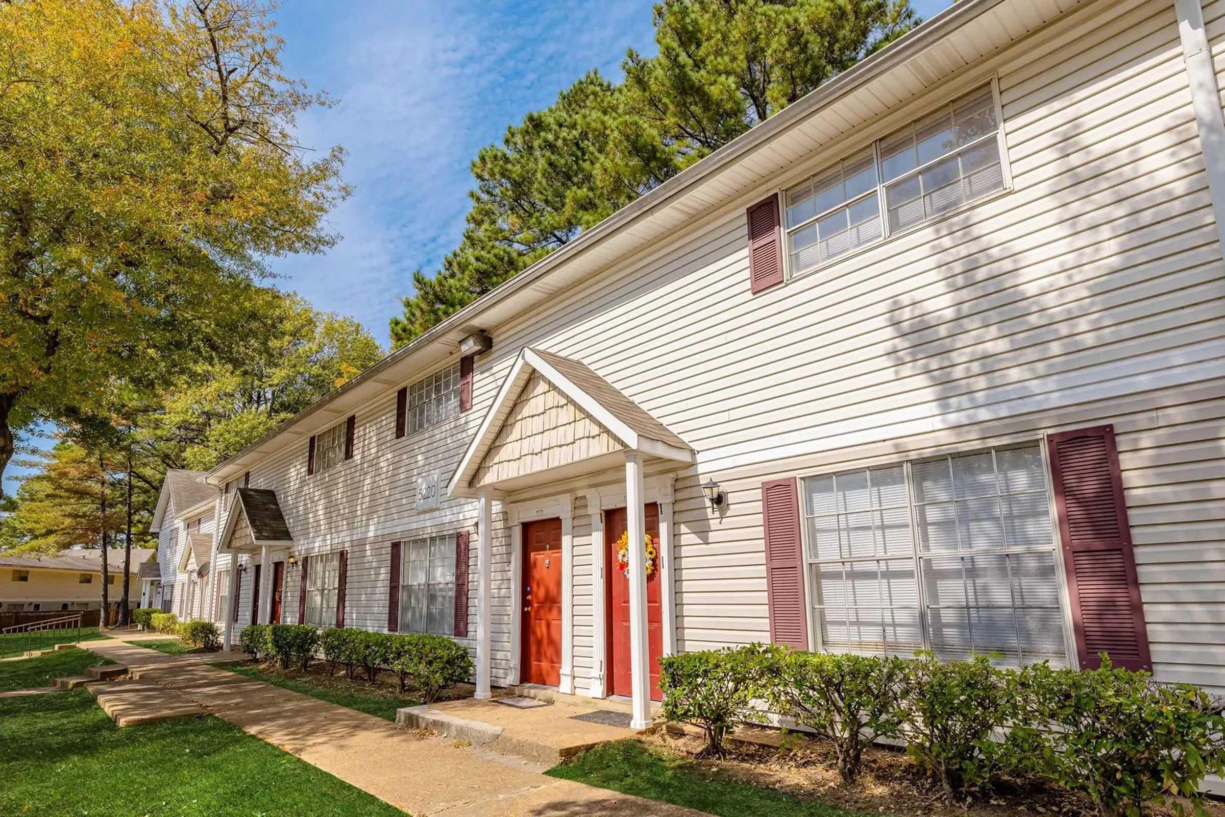 A two-story apartment building with a light-colored exterior, featuring multiple windows and red front doors. A well-maintained pathway leads to the entrance, surrounded by manicured shrubs and trees. The sky is partly cloudy, indicating a pleasant day.