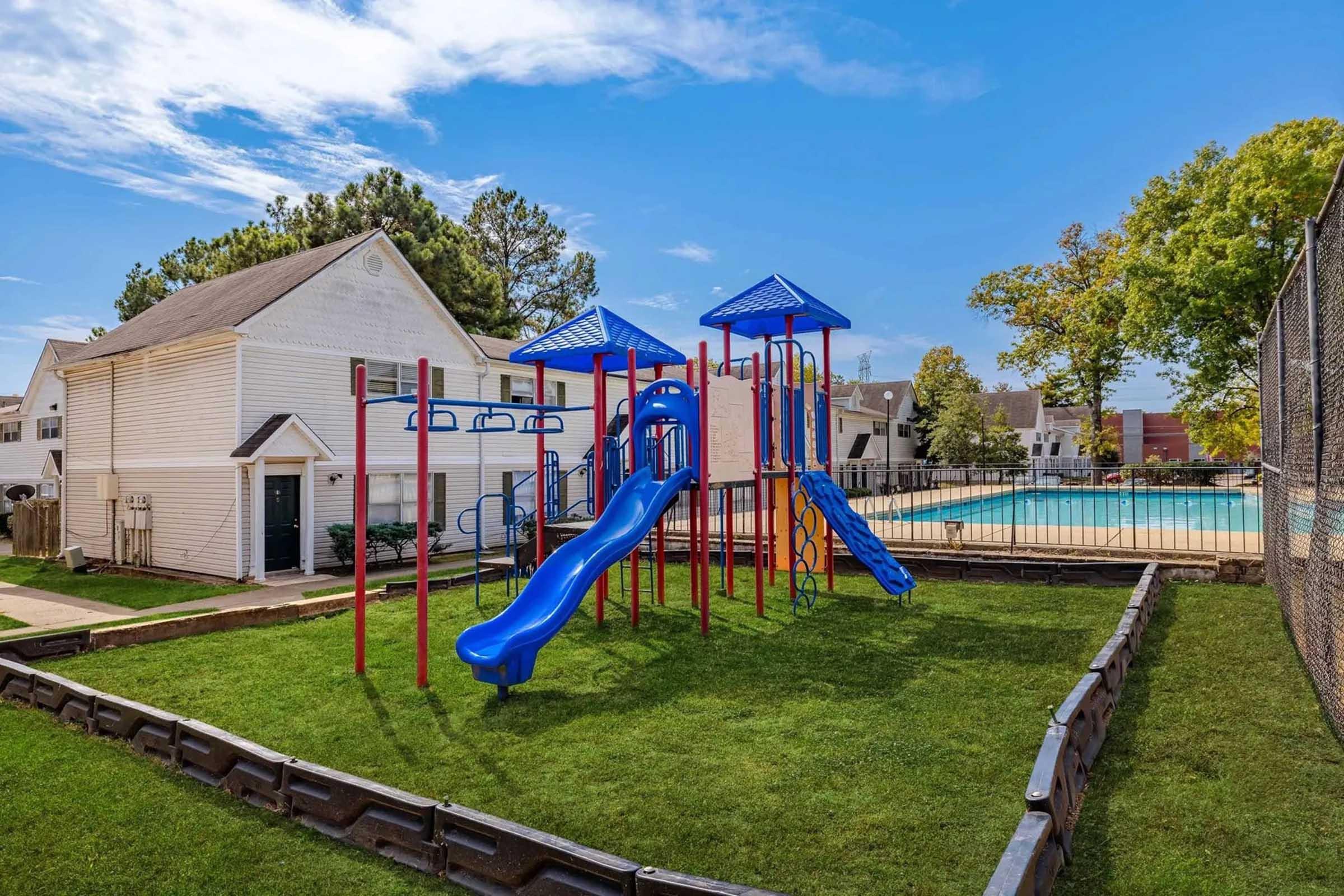 A colorful playground with blue slides and climbing structures on a grassy area, adjacent to a swimming pool. In the background, there are residential buildings and trees under a clear blue sky with some clouds. The area is enclosed with a fence.