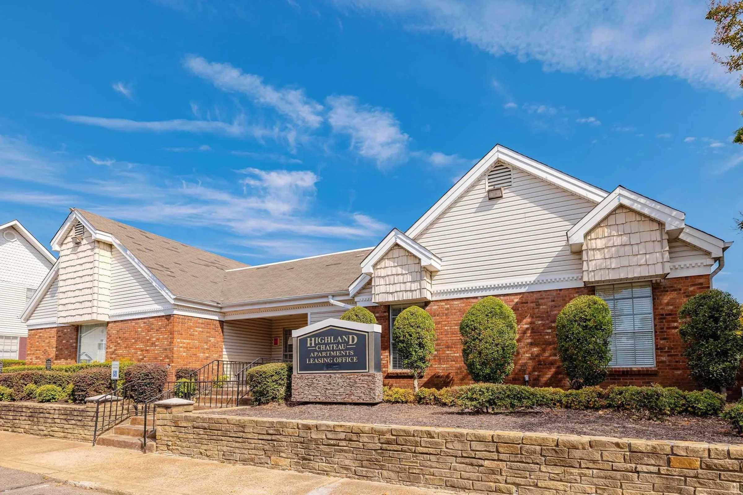 Brick building with white siding and a peaked roof, featuring a sign that reads "Highland Apartments Leasing Office." The landscaping includes trimmed bushes and a clear blue sky in the background. The entrance has steps leading up to the front door.