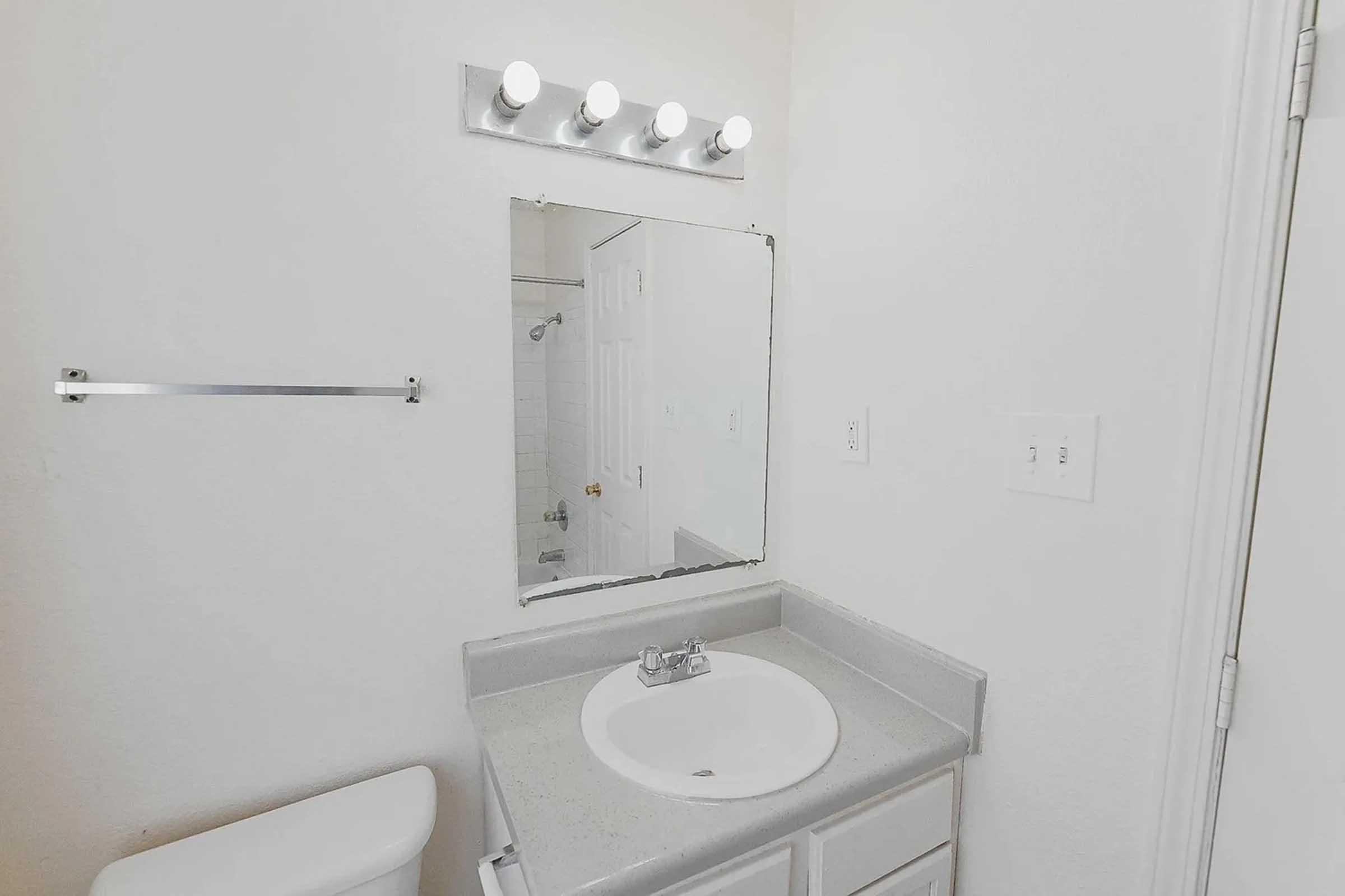 A clean and simple bathroom featuring a white sink with a circular basin, a large mirror above the sink, and four light bulbs mounted on a fixture. The walls are painted white, and a towel rack is visible beside the mirror. The bathroom has a neutral and bright appearance.