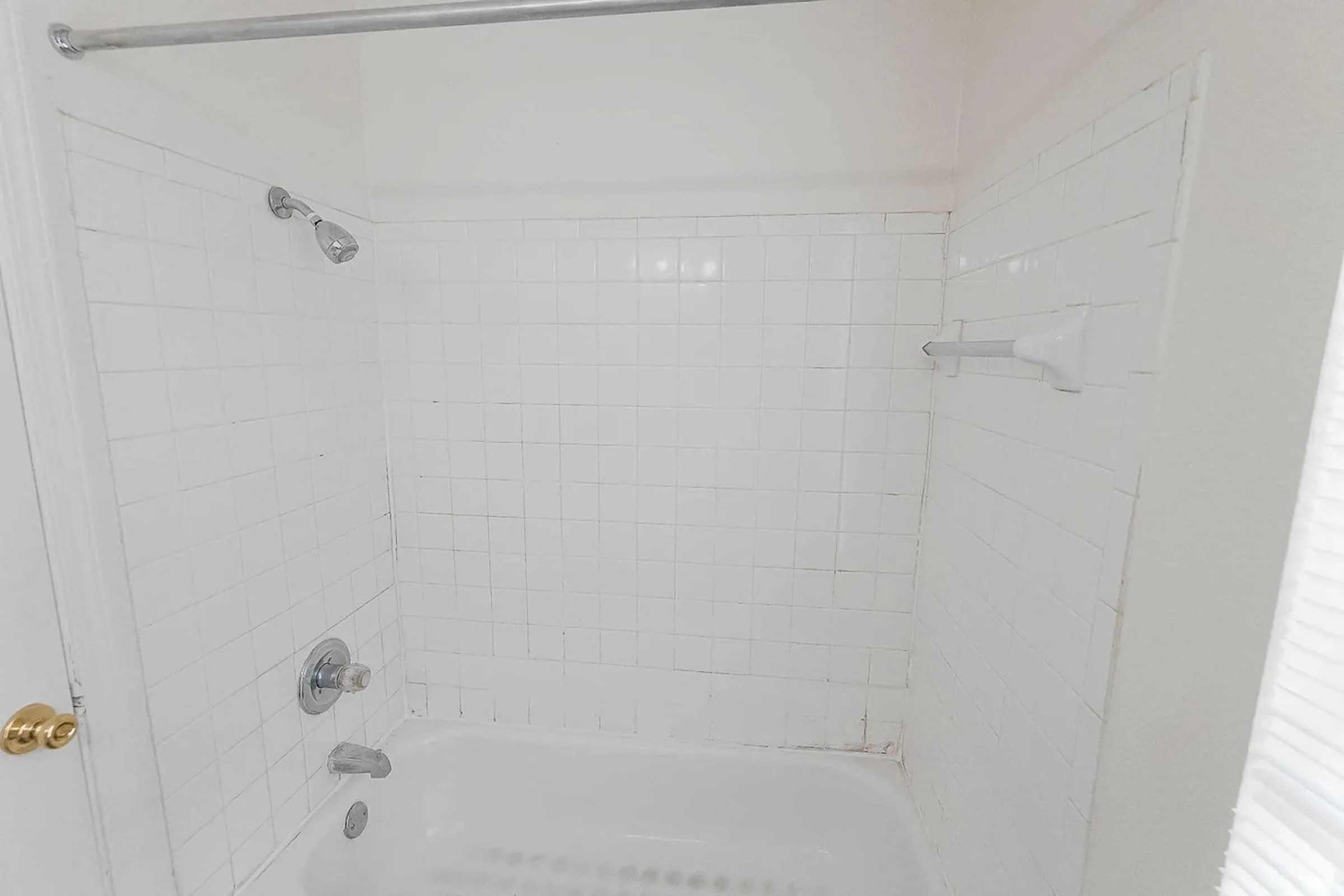A clean, white tiled shower area featuring a showerhead, a faucet, and a soap dish. The shower is empty with bright lighting, showcasing the simplicity of the design. The tiles are arranged in a grid pattern, and the overall aesthetic is minimalistic and functional.