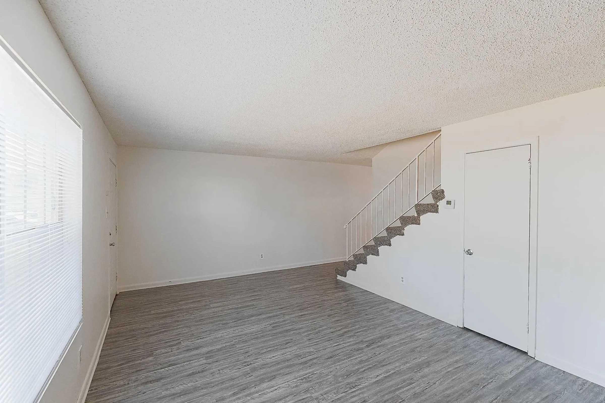 A spacious, empty living area featuring light-colored walls and a textured ceiling. A staircase with white railing is visible in the background. The floor is covered with light wood-like laminate, and there are large windows allowing natural light to enter the room. A closed door is on the right side.