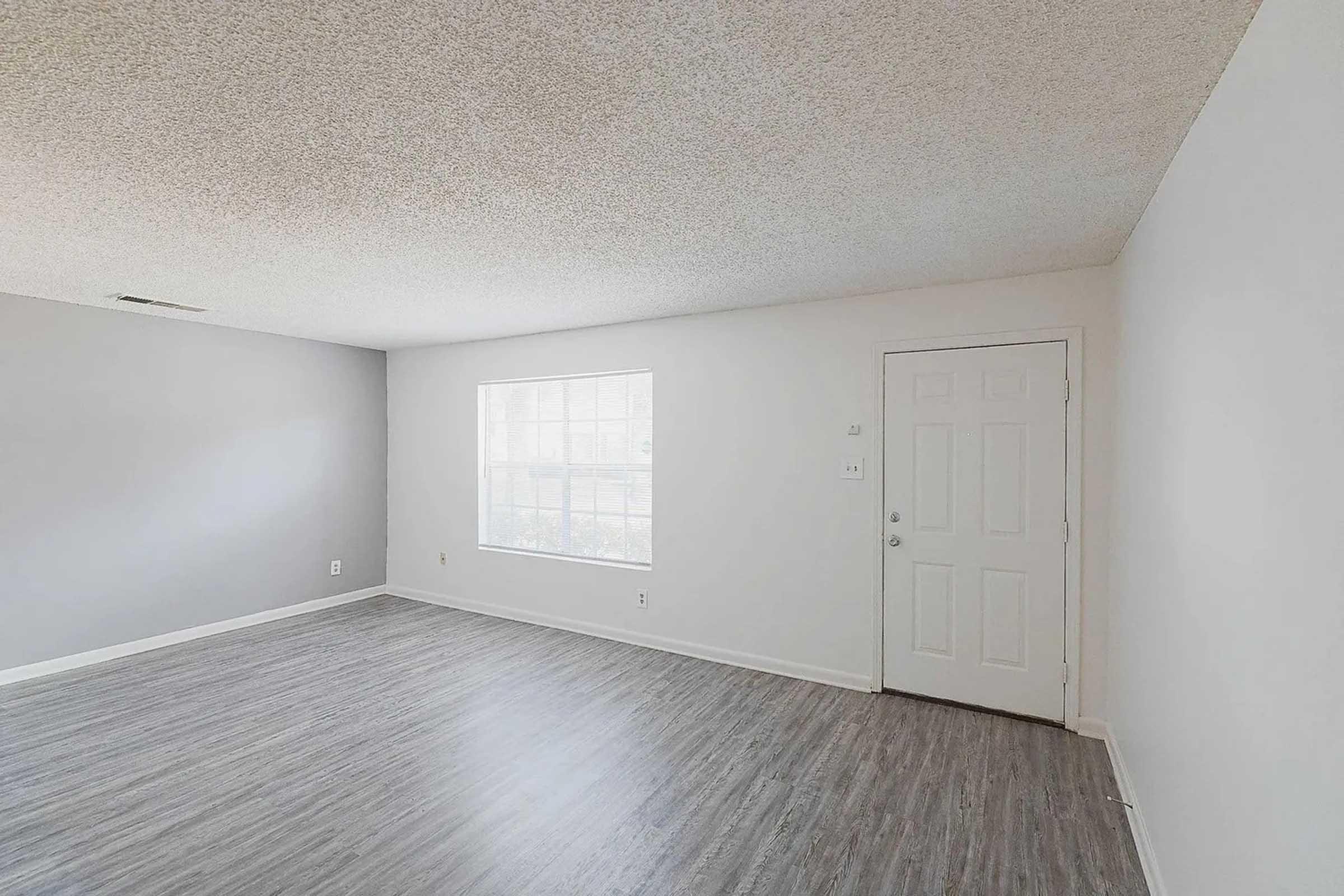 A spacious, empty room with light gray walls and a textured ceiling. A large window on the left allows natural light to enter, illuminating the light-colored flooring. A white door is in the corner, leading outside. The overall atmosphere is clean and uncluttered, perfect for interior design.
