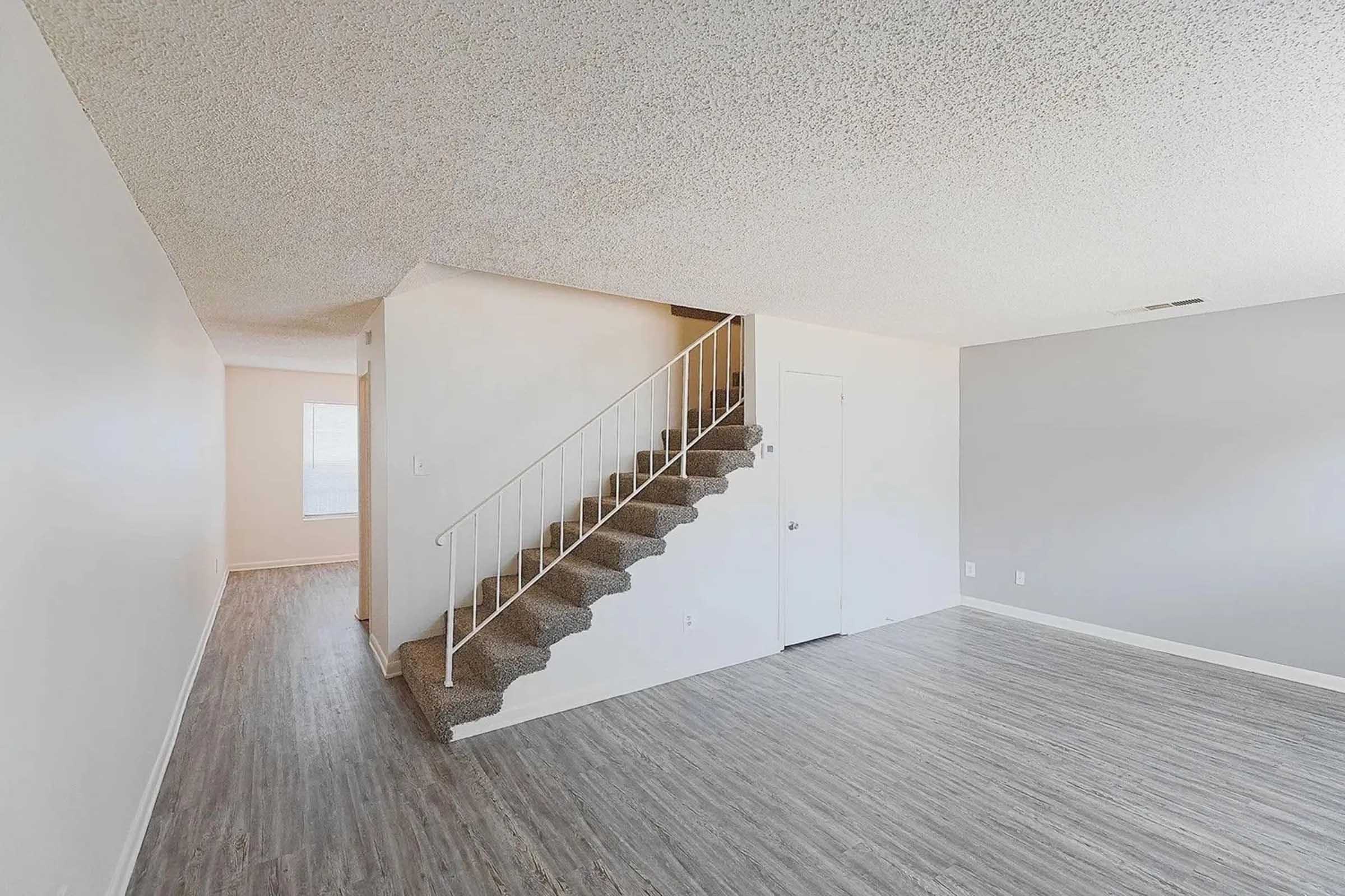 Interior view of a spacious living area featuring light-colored walls, a staircase leading to an upper level, and modern flooring. The room appears well-lit with natural light coming from nearby windows, creating an inviting atmosphere.