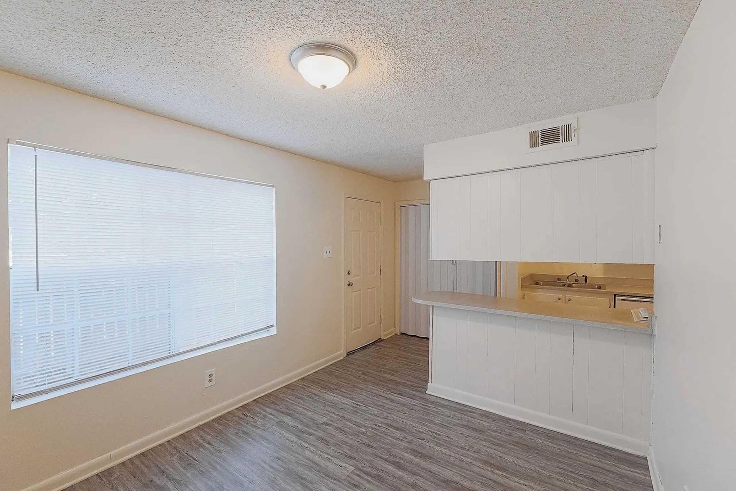 A well-lit, unfurnished living space featuring a large window with blinds, a door leading to the hallway, and a small kitchen area with a counter. The walls are painted a light color and the floor is a light wood laminate. The ceiling has a simple light fixture.