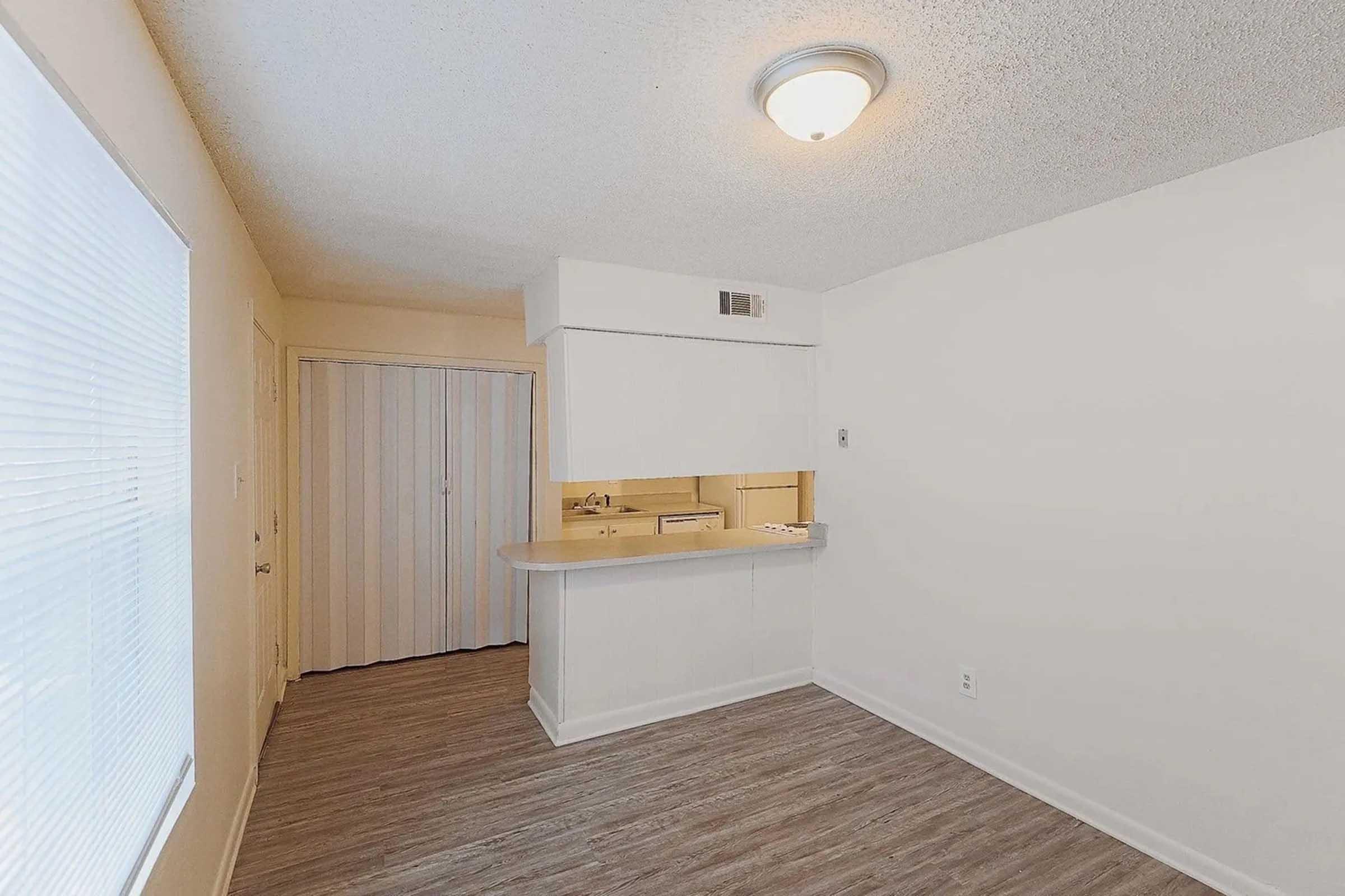A small, empty room with light-colored walls and a textured ceiling. The floor is covered in light wood laminate. To the left, there is a window with sheer blinds, and in the background, a small kitchen area is visible with white cabinetry. A door leads to another room, and there is a ceiling light fixture.