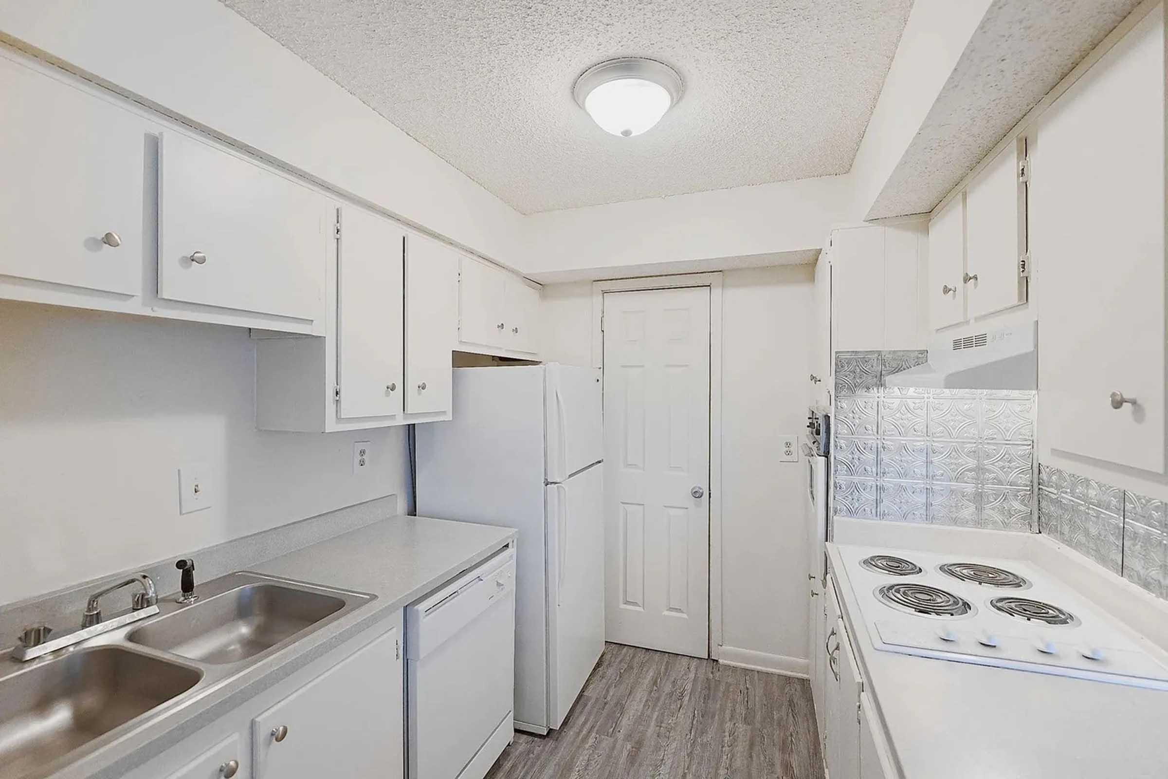 A compact kitchen featuring white cabinets, a double sink, a dishwasher, and a refrigerator. The counter is clean with a stove top, and the walls have a neutral color. A door leads to another room, and the ceiling has a round light fixture. The flooring is a light wood-like material.
