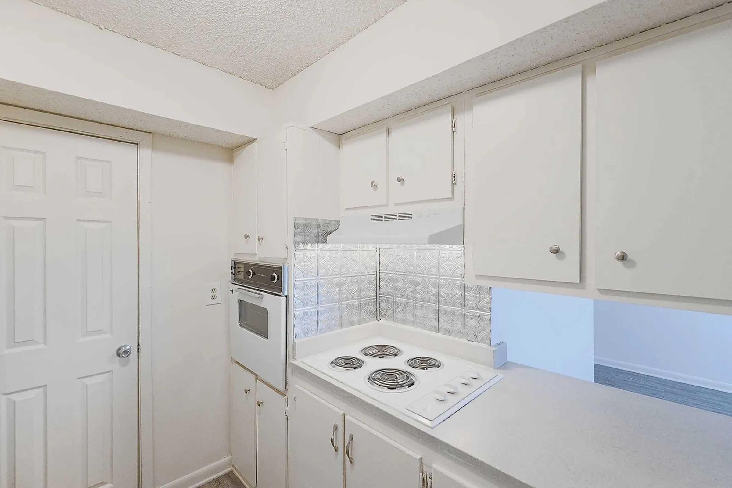 A small kitchen featuring white cabinets, a countertop with a stovetop, and an oven. The backsplash has a shiny, reflective surface. A closed door is visible on the left side, and the space appears well-lit and tidy.