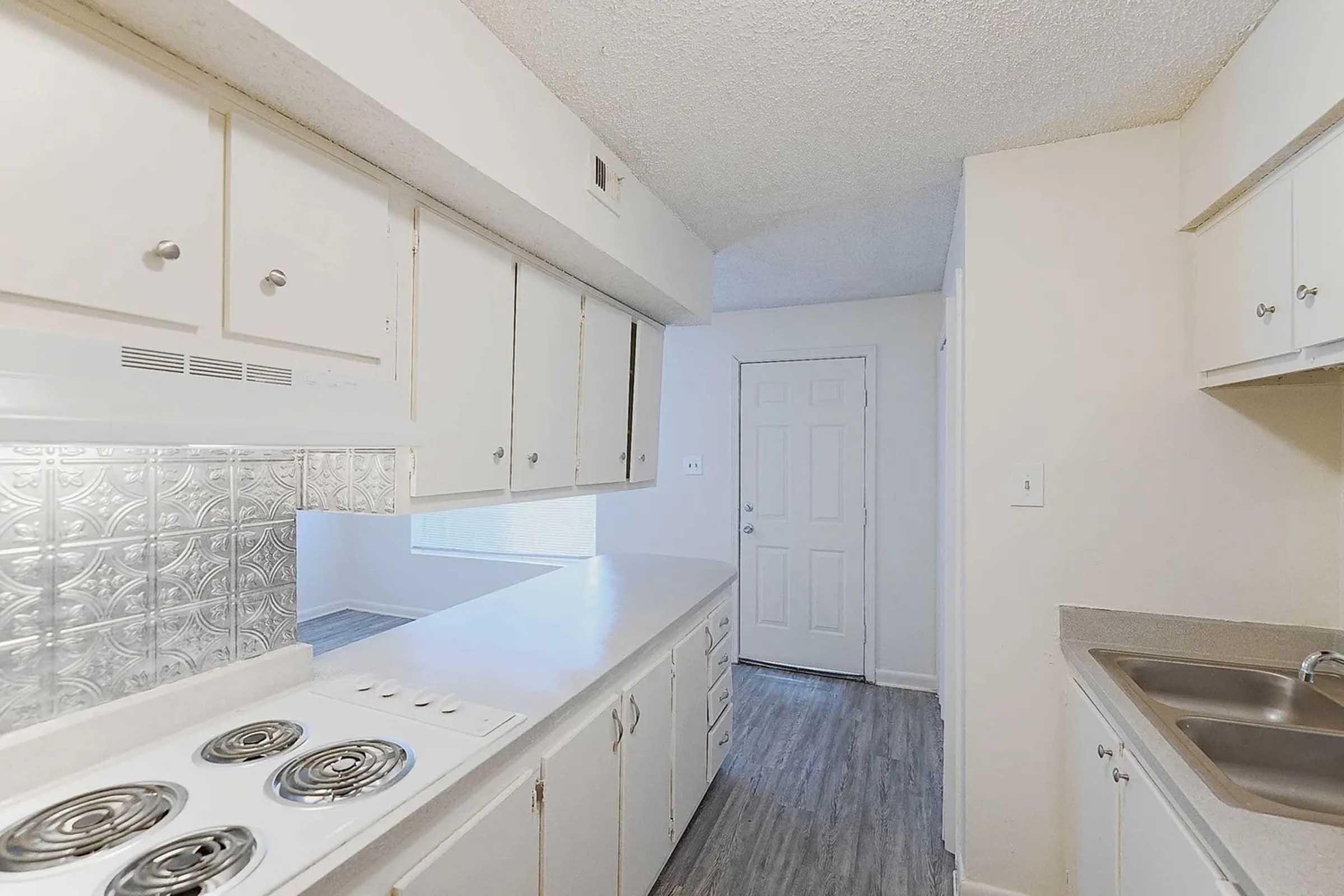 A spacious kitchen featuring white cabinetry, a stovetop with burners, a stainless steel sink, and a white countertop. Natural light enters through a window, and there is an entrance door visible at the end of the hallway. The flooring has a light wood-like finish, providing a clean, bright atmosphere.