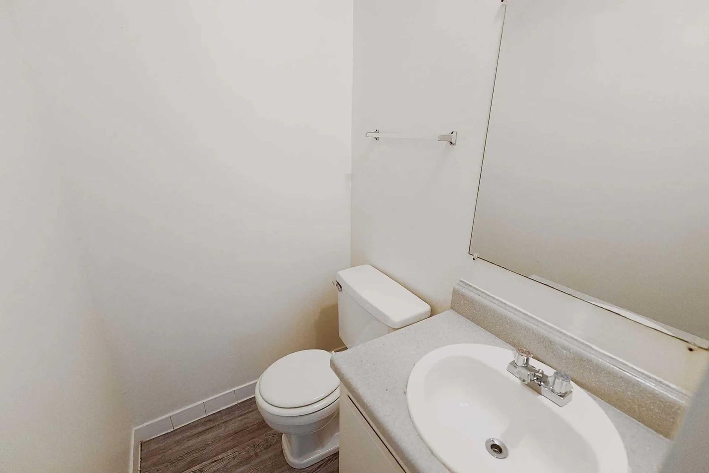 A simple bathroom featuring a white toilet, sink with a mirror above, and a light-colored wall. The floor has a wooden appearance, and there's a towel bar on the wall. The space is well-lit and minimalistic.