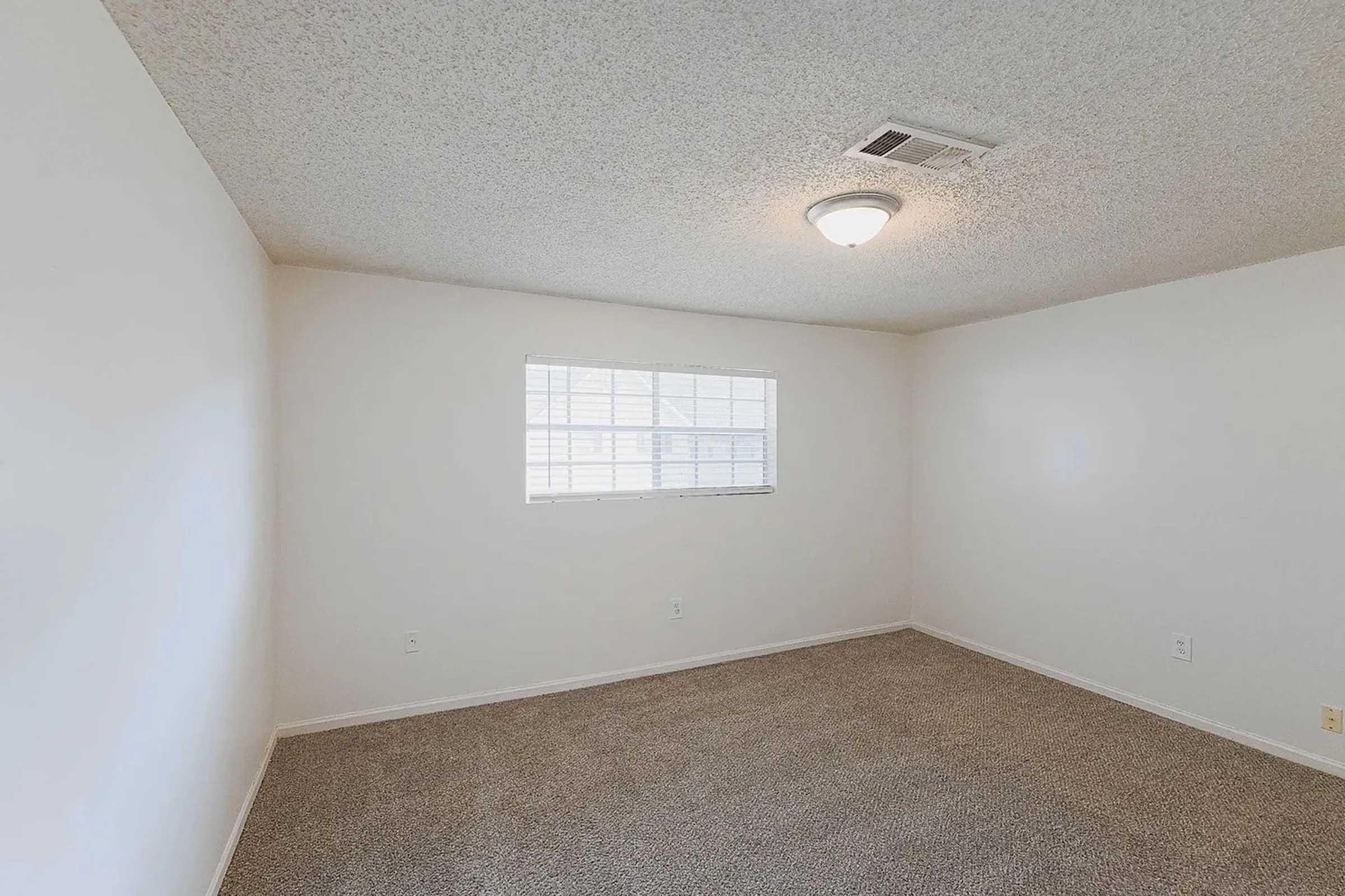 Empty room with light-colored walls and a textured ceiling. A small window allows natural light to enter, and the floor is covered with beige carpet. The overall feel is spacious and clean, with no furniture or decorations present.