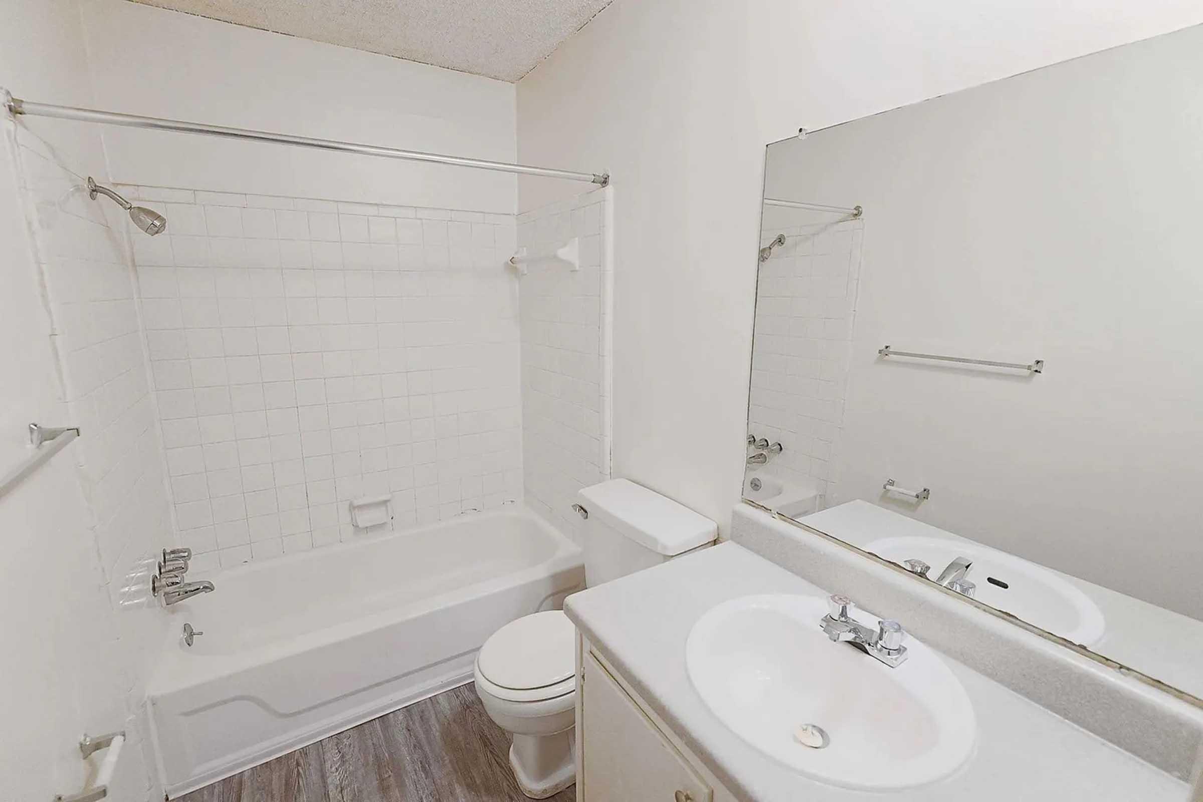 A clean and simple bathroom featuring a bathtub with shower, a toilet, and a sink with a mirror above it. The walls are tiled, and the floors have a wooden appearance. The overall color scheme is light and neutral, creating a spacious feel.