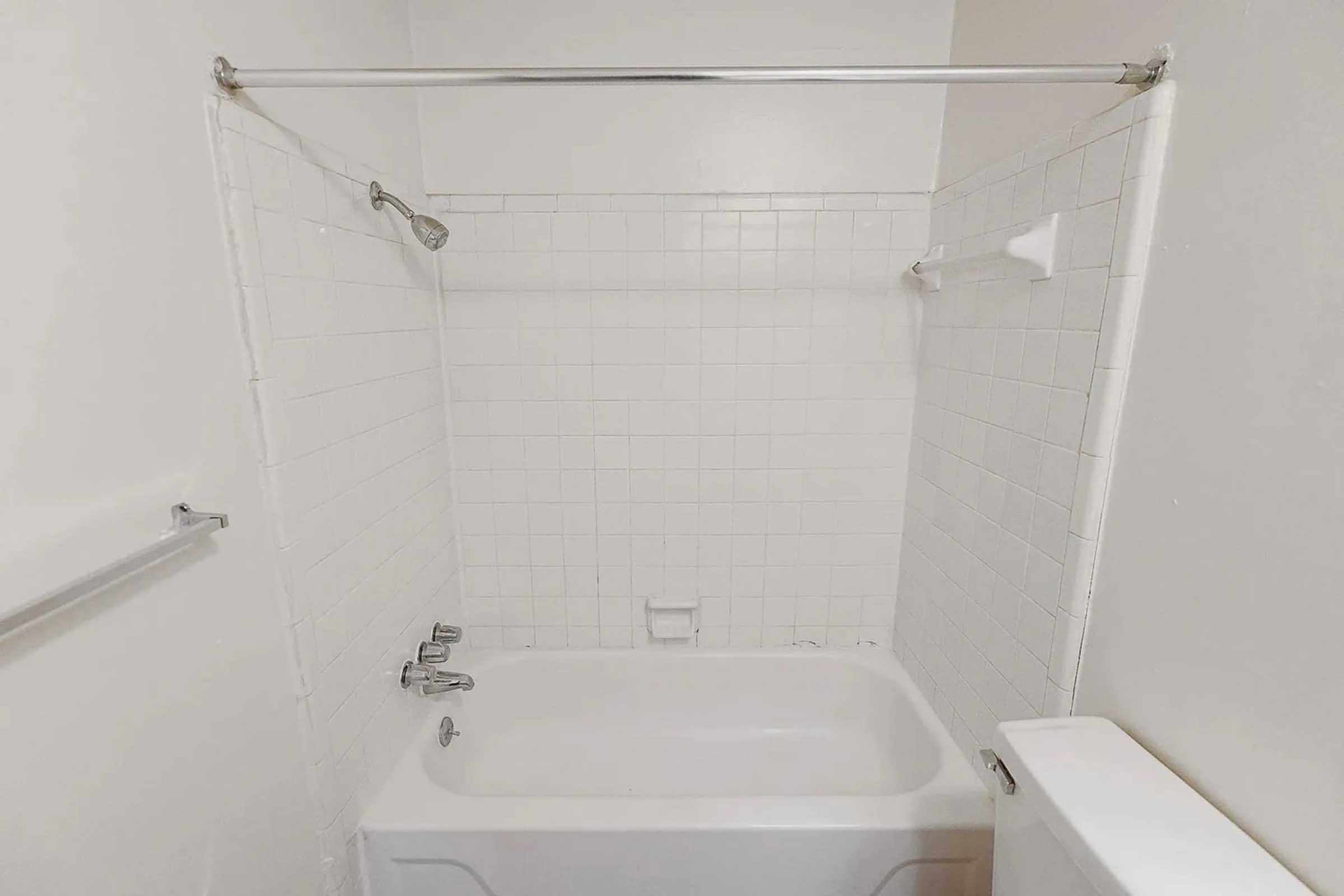 A clean, white tiled bathroom featuring a bathtub with a showerhead attached to a chrome rod. There's a wall-mounted towel bar on the left and a toilet visible on the right, all in a minimalist, bright space.