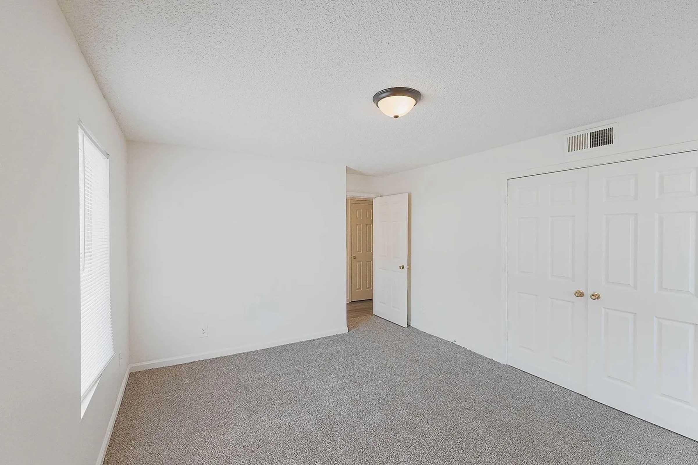 A spacious, empty room featuring light-colored walls, textured ceiling, and carpeted flooring. There is a window on the left side, allowing natural light. A doorway leads to another room, and white double closet doors are visible on the right. The overall ambiance is clean and bright, suitable for various uses.