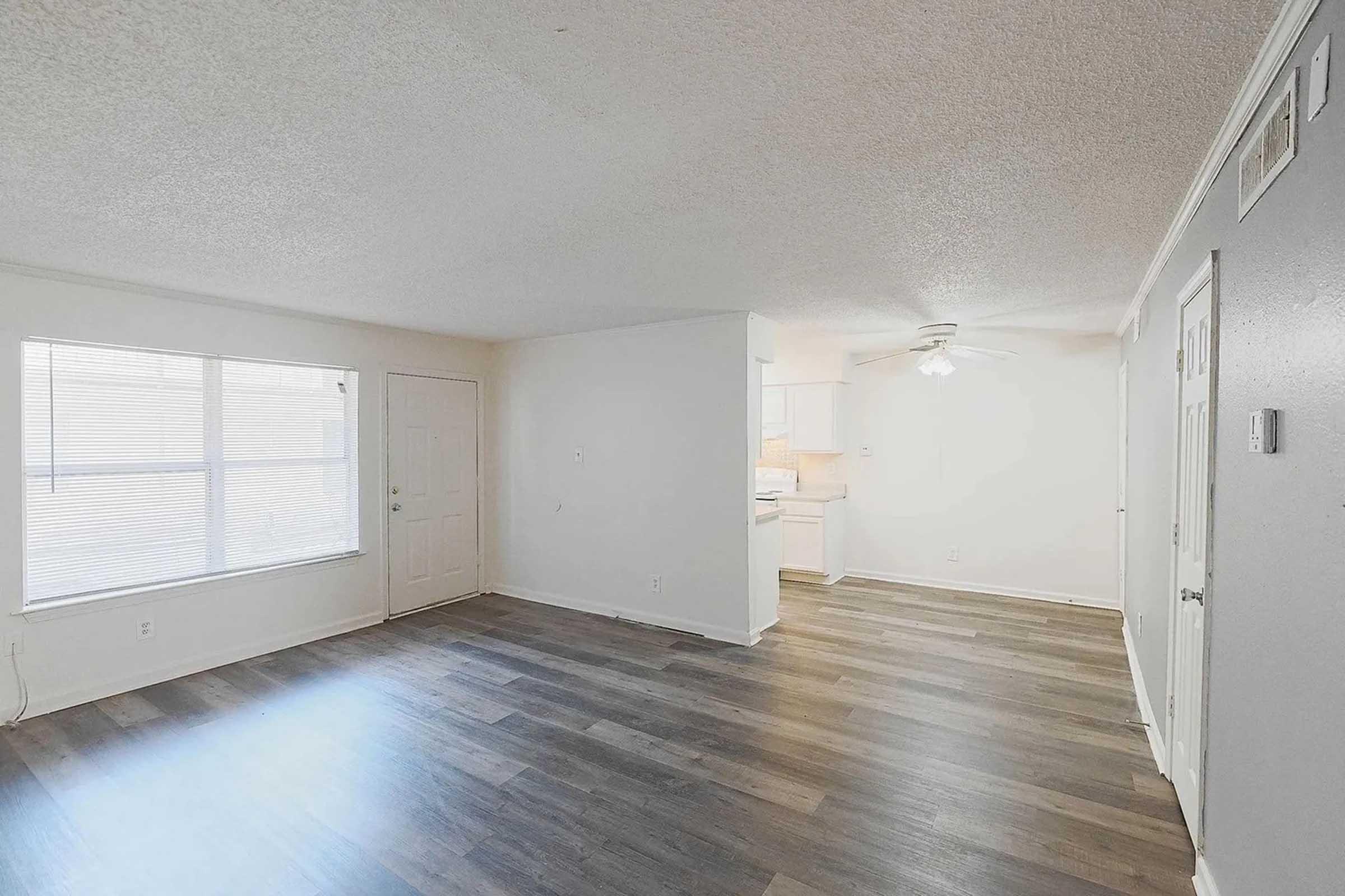 A spacious, well-lit living area with light-colored walls and new vinyl flooring. It features a window with blinds, a front door, and a view of a kitchen area in the background. The ceiling is fitted with a fan, and there's ample natural light entering the room.