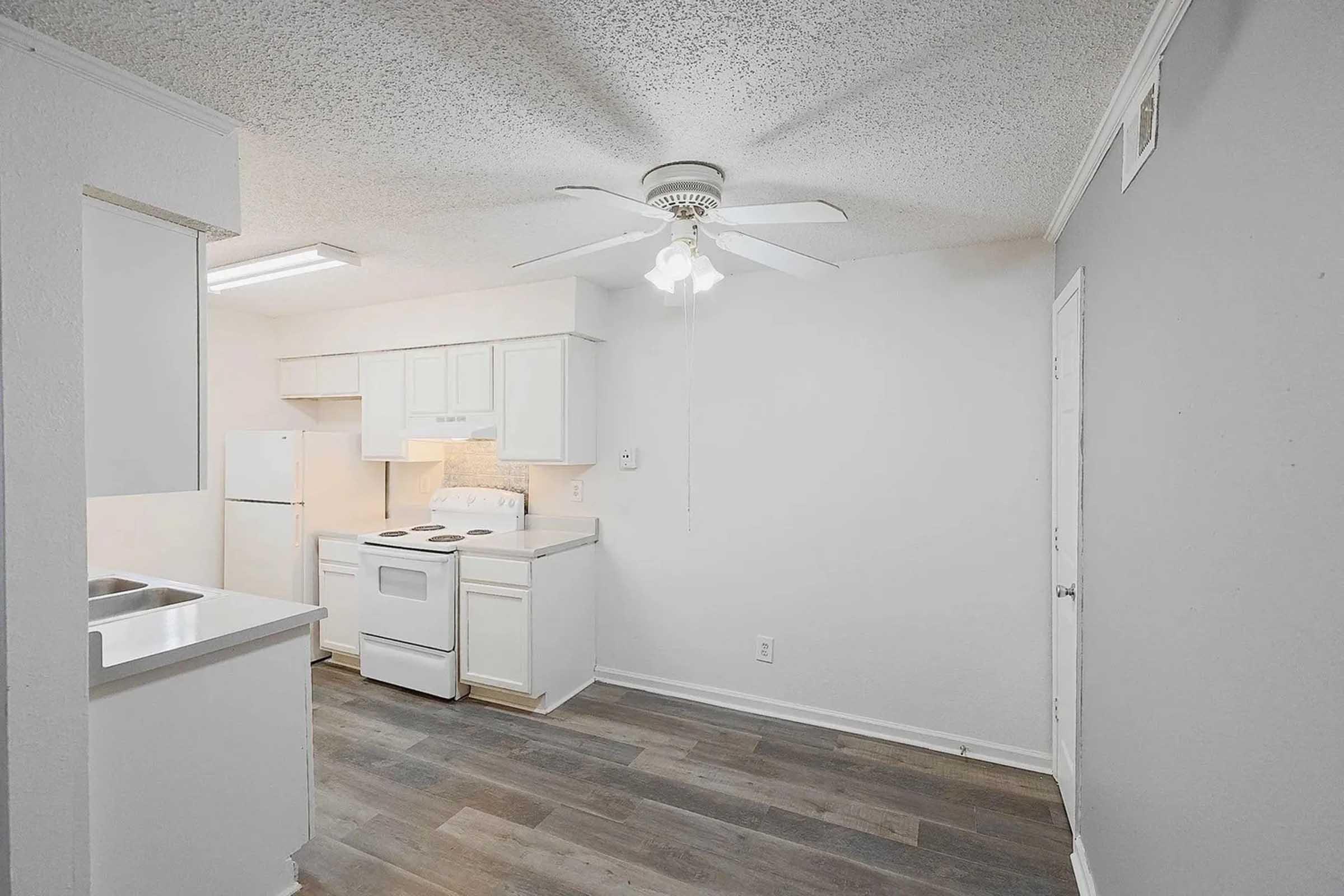 A small, modern kitchen featuring white cabinetry, a ceiling fan, and appliances including a stove, refrigerator, and sink. The flooring is wood-like, and the walls are painted light gray. Bright overhead lighting enhances the inviting atmosphere.