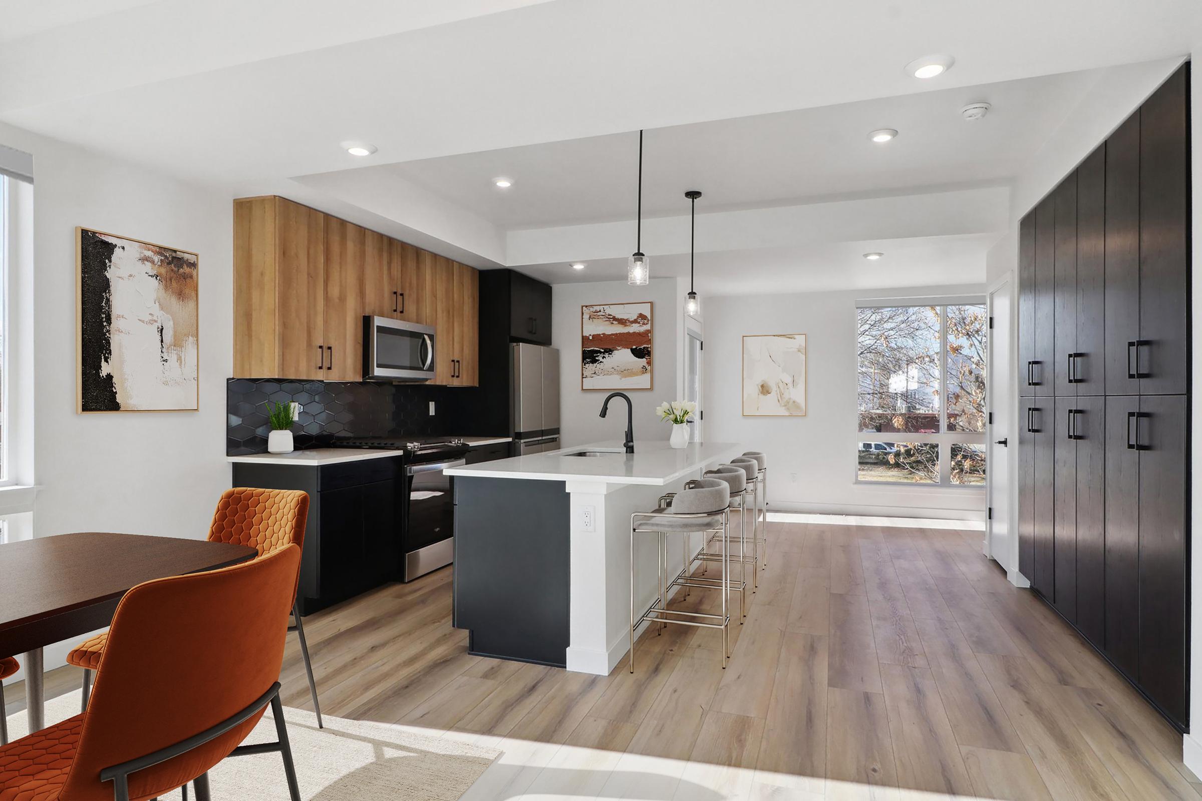 A modern kitchen and dining area featuring sleek black and wood cabinetry, stainless steel appliances, and a large island with bar stools. Bright windows allow natural light, highlighting decorative artwork on the walls. A dining table with stylish orange chairs complements the contemporary design.