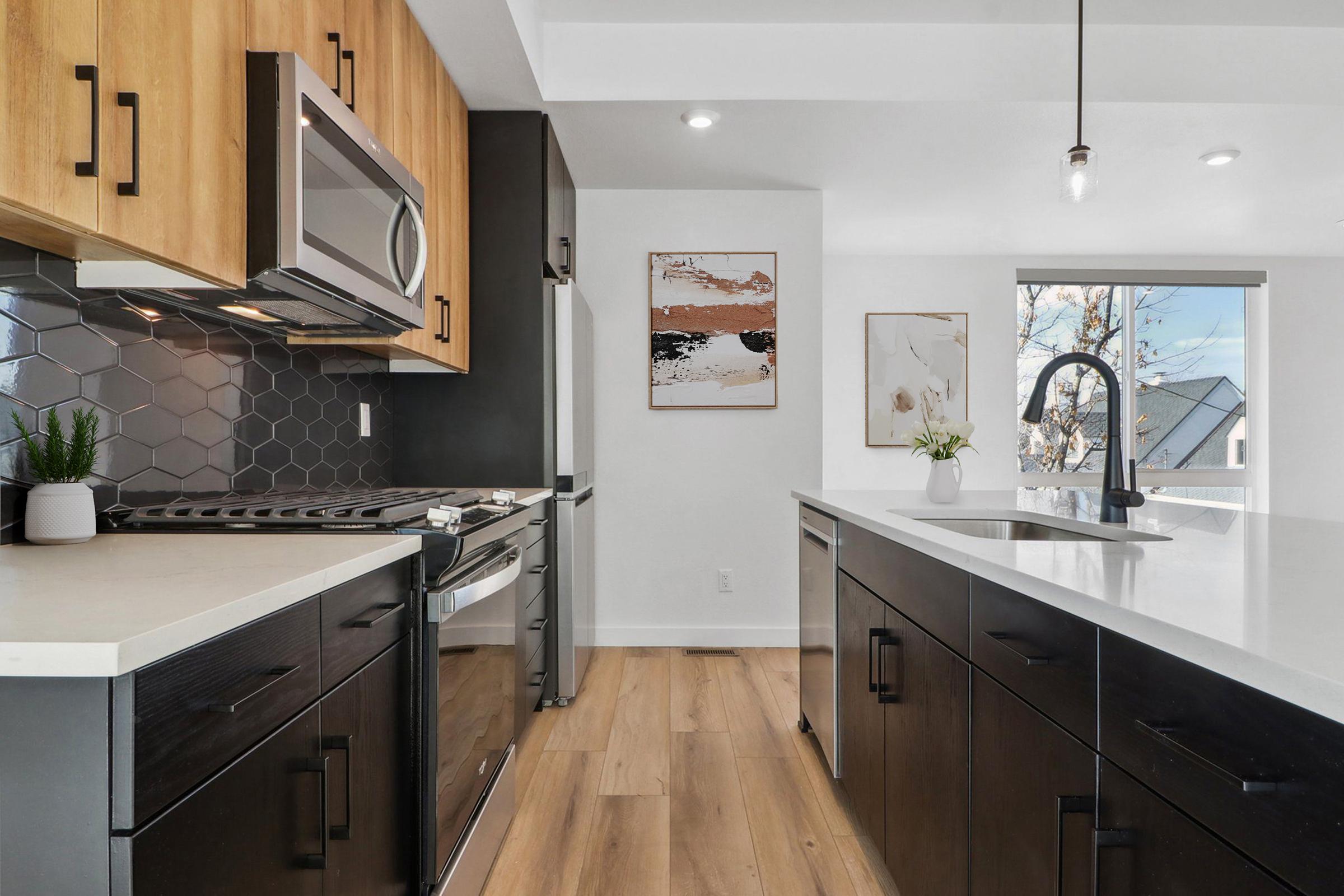 Modern kitchen featuring a mix of black and wood cabinetry, stainless steel appliances, and a stylish countertop. The backsplash is made of hexagonal tiles, and there's a large window with a view. Decor includes a small plant and two framed artworks on the wall. Natural light fills the space, highlighting the clean design.