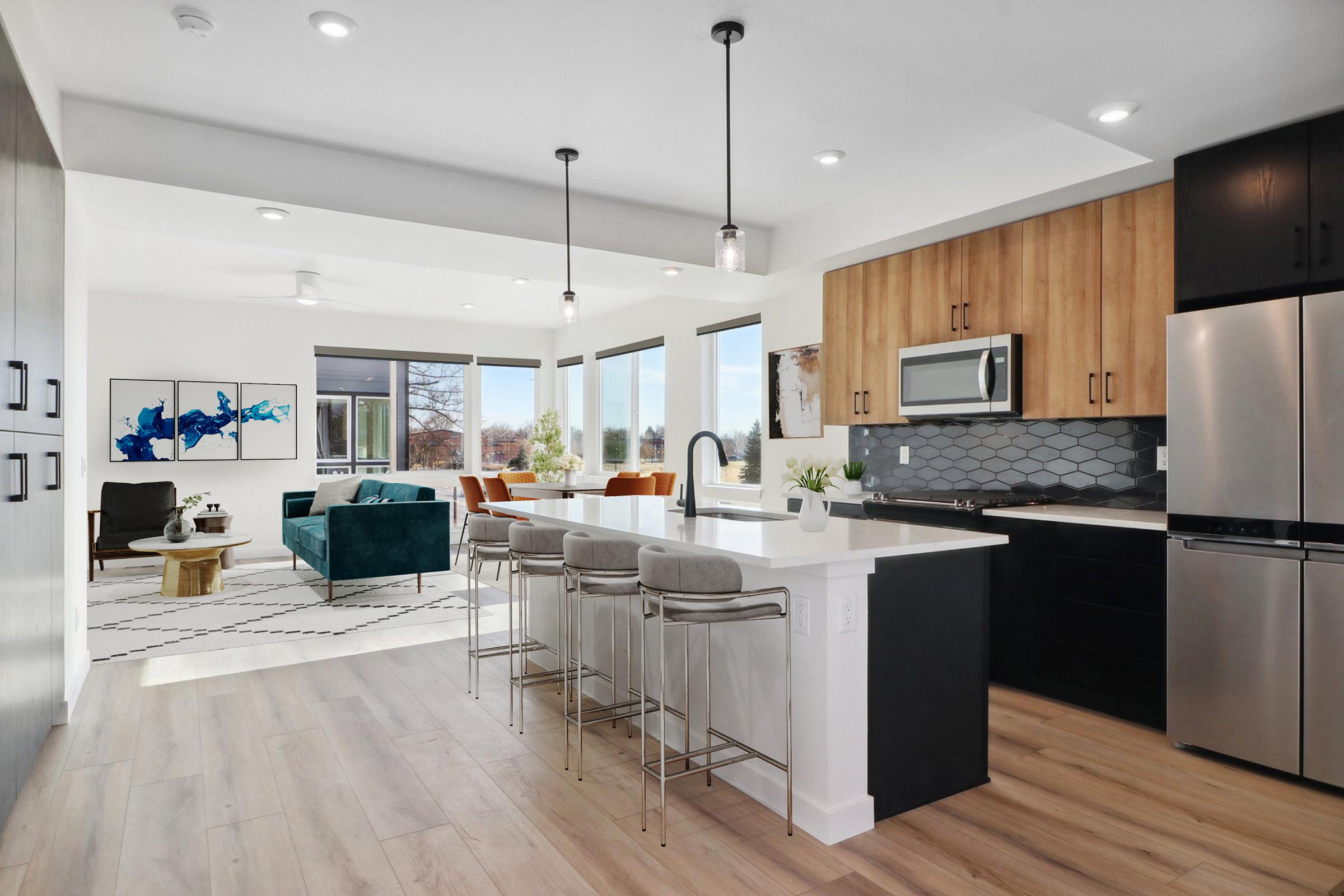 Modern kitchen with light wooden cabinets and dark cabinetry, featuring a large island with bar stools. The open space connects to a living area with green velvet chairs, large windows providing natural light, and a stylish rug on hardwood flooring. Decorative artwork is displayed on the wall.