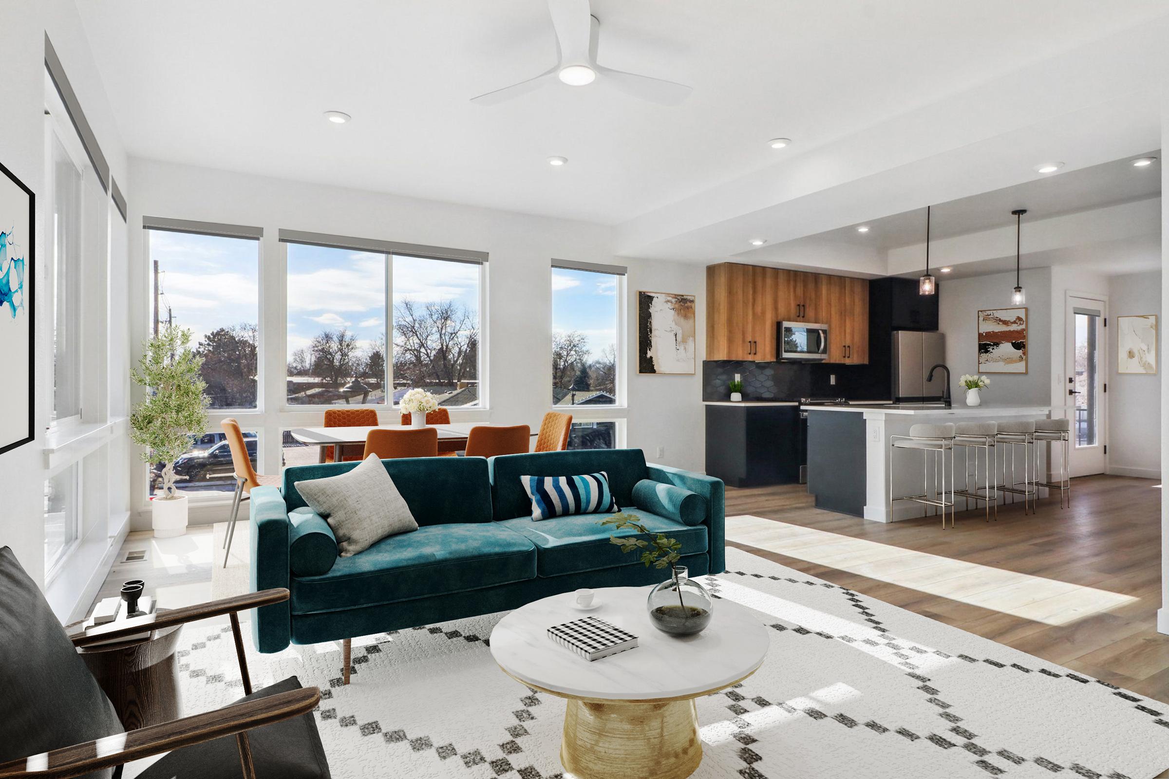 Bright and modern living space featuring a cozy teal sofa with pillows, a round coffee table, and a stylish area rug. The open layout includes a dining area with a wood table and chairs, a sleek kitchen with dark cabinetry and stainless steel appliances, and large windows allowing natural light to fill the room.