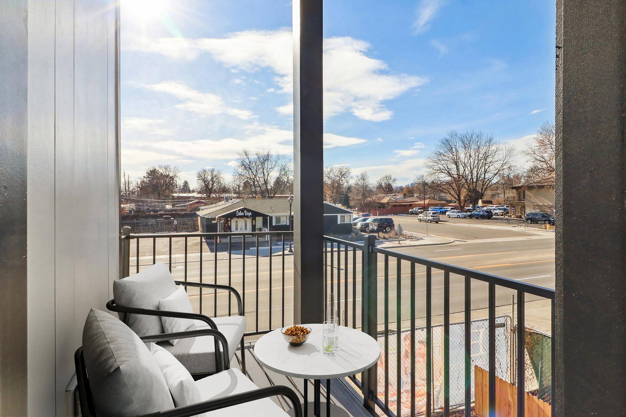 A balcony view featuring two cozy chairs and a small round table. On the table, there are snacks and glasses. In the background, there's a street scene with parked cars and trees, under a bright blue sky with a few clouds. Sunlight illuminates the space, creating a relaxed atmosphere.