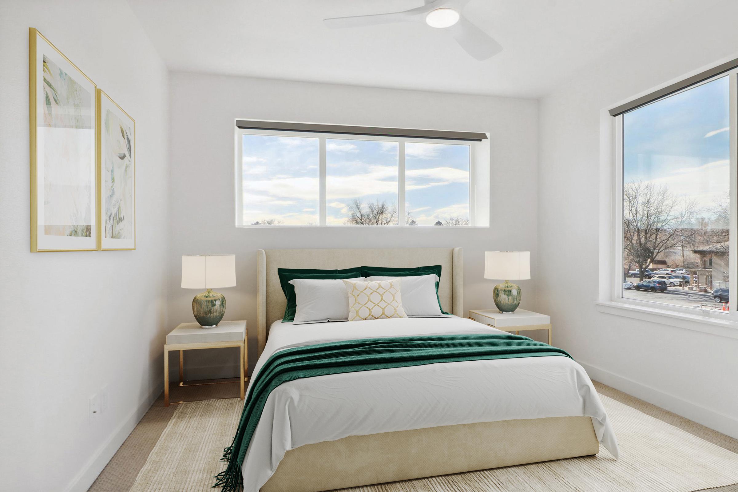A stylish bedroom featuring a queen-sized bed with a white and green bedspread. Flanking the bed are two modern nightstands with table lamps. Large windows allow natural light to fill the room, showcasing a view of trees outside. The walls are painted white, and framed artwork is displayed above the bed.