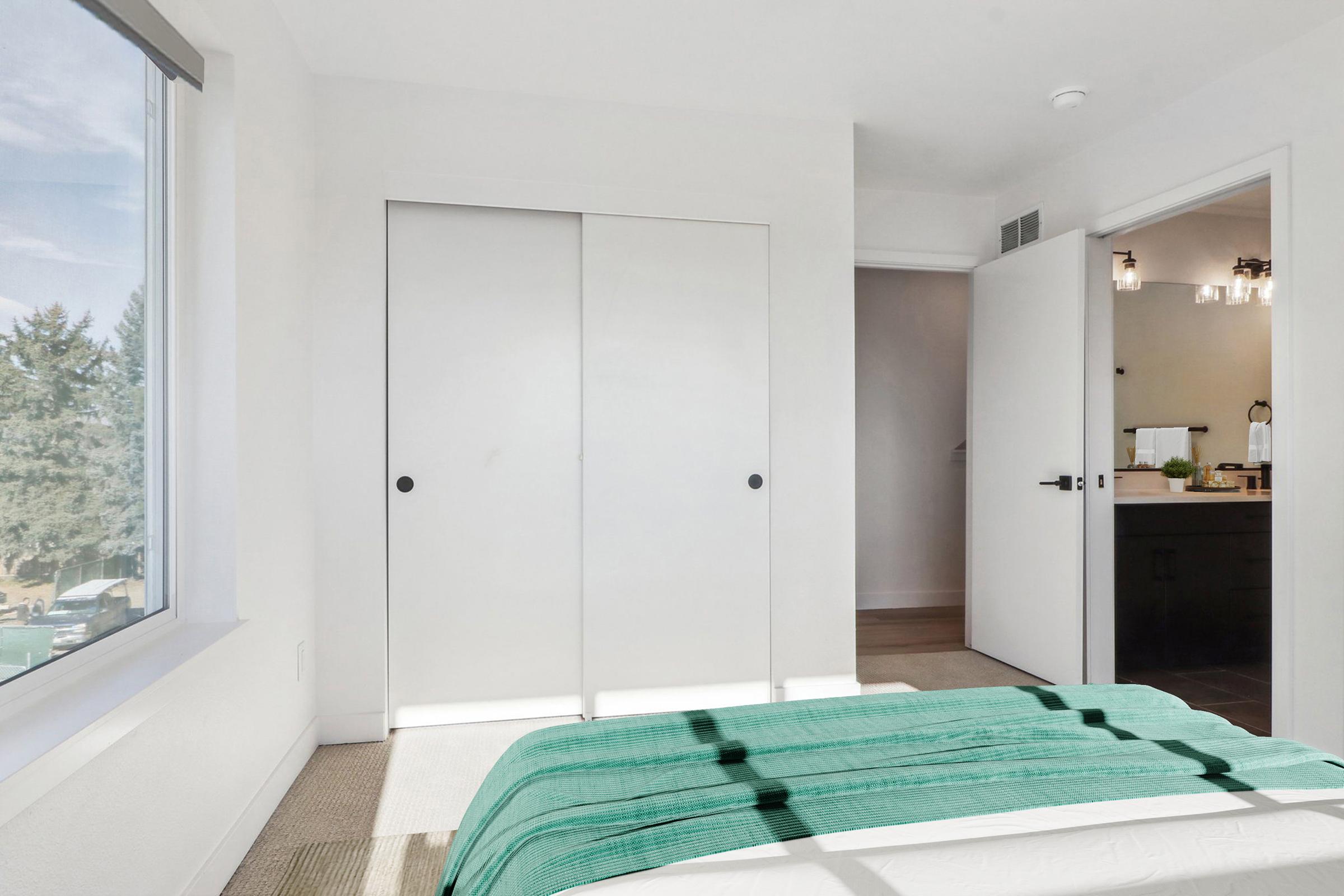 A bright, modern bedroom featuring a neatly made bed with a teal throw blanket. There are white walls and closet doors on one side, while a doorway leads to a bathroom. Natural light streams in through a large window, enhancing the airy feel of the space.