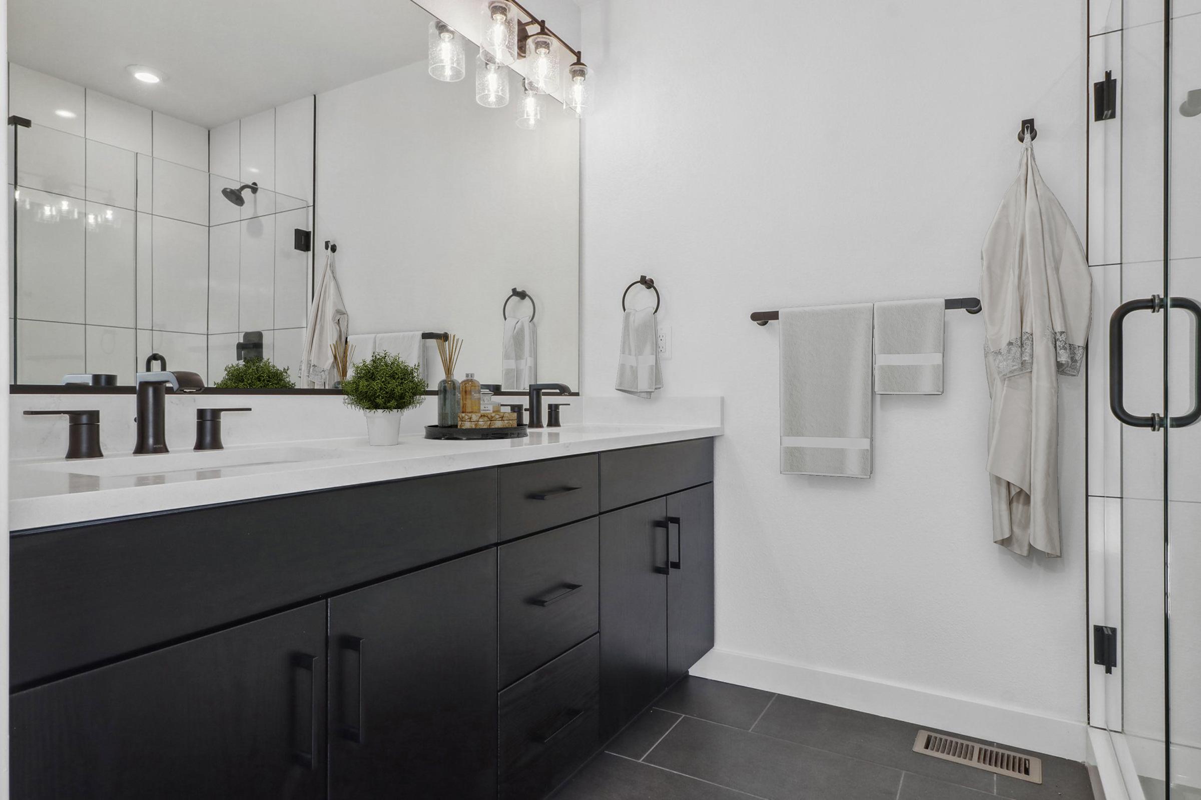 A modern bathroom featuring sleek black cabinetry, a spacious white countertop, and a large mirror. The decor includes stylish lighting fixtures and neatly arranged towels. A small plant adds a touch of greenery to the minimalist design, while the overall aesthetic is contemporary and monochromatic.