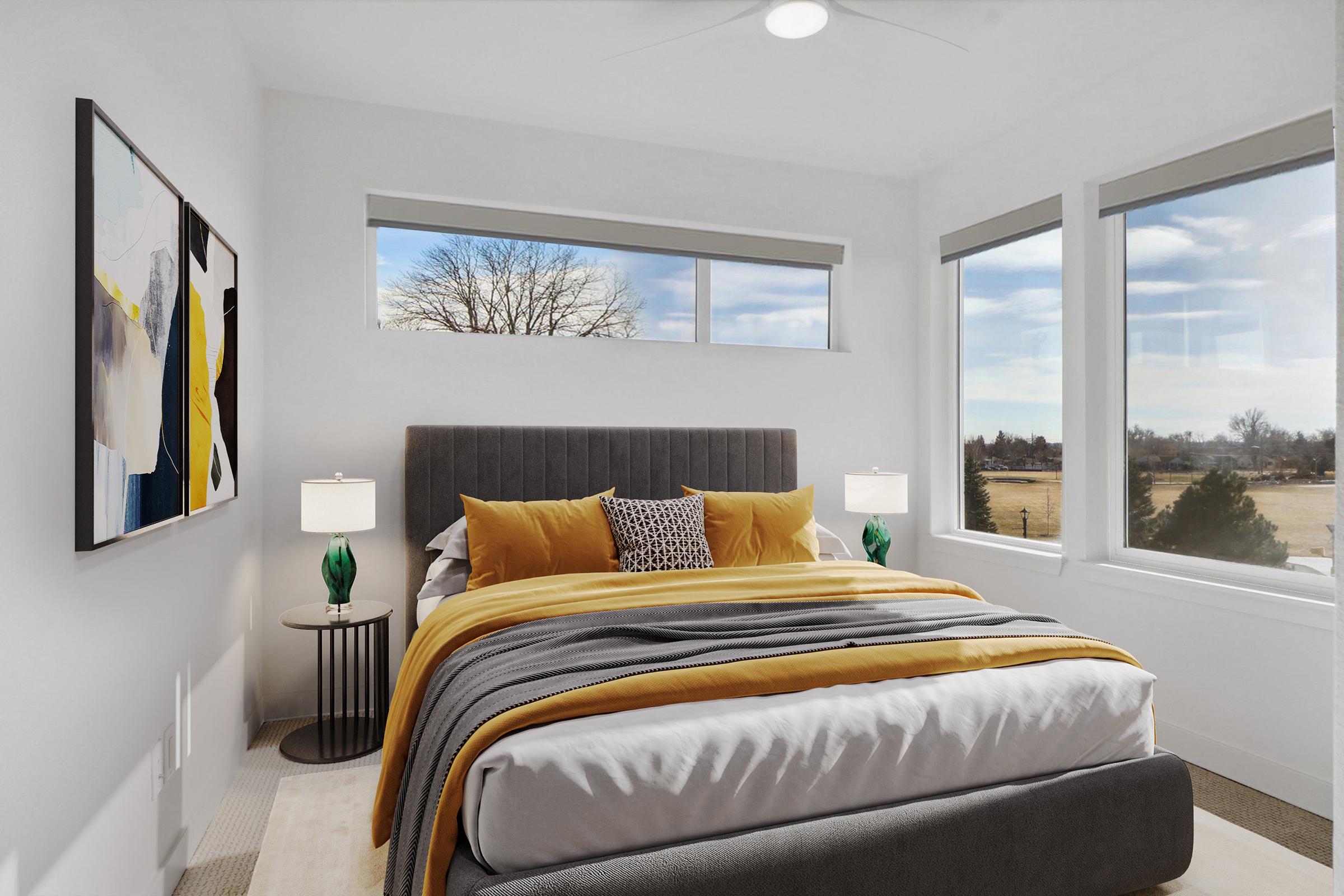 A modern bedroom with a large bed featuring gray and yellow linens, accented by decorative pillows. Two bedside tables with green lamp accents flank the bed. The room has large windows with a view of trees and open fields, and colorful artwork is displayed on the wall. The overall atmosphere is bright and inviting.