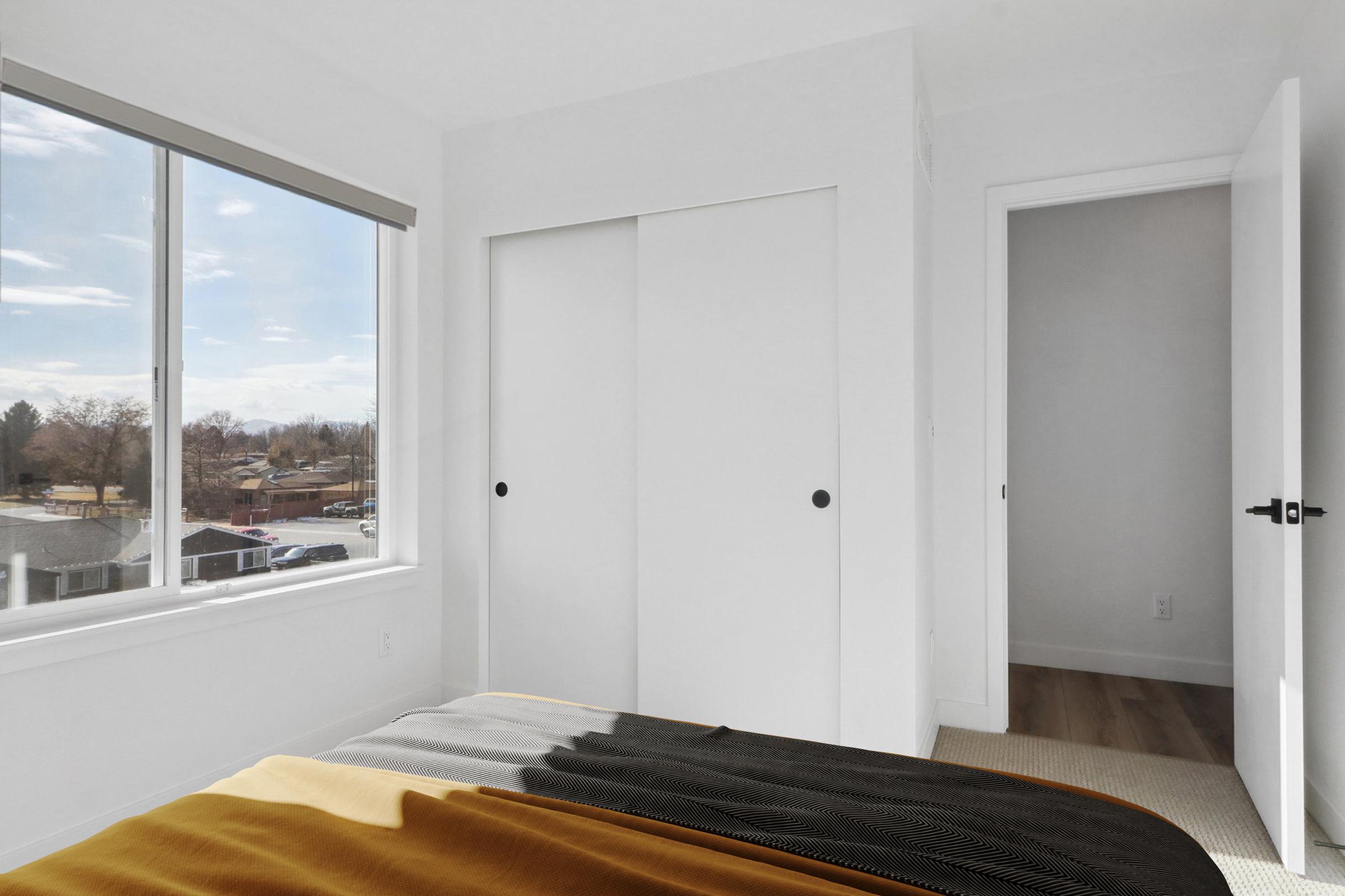 A bright and modern bedroom featuring a large window with a view of the outdoors, a neatly made bed with a black and gold blanket, and a closet with sliding doors. The room has light-colored walls and floor, creating a spacious and airy feel. A partially open door leads to another room.