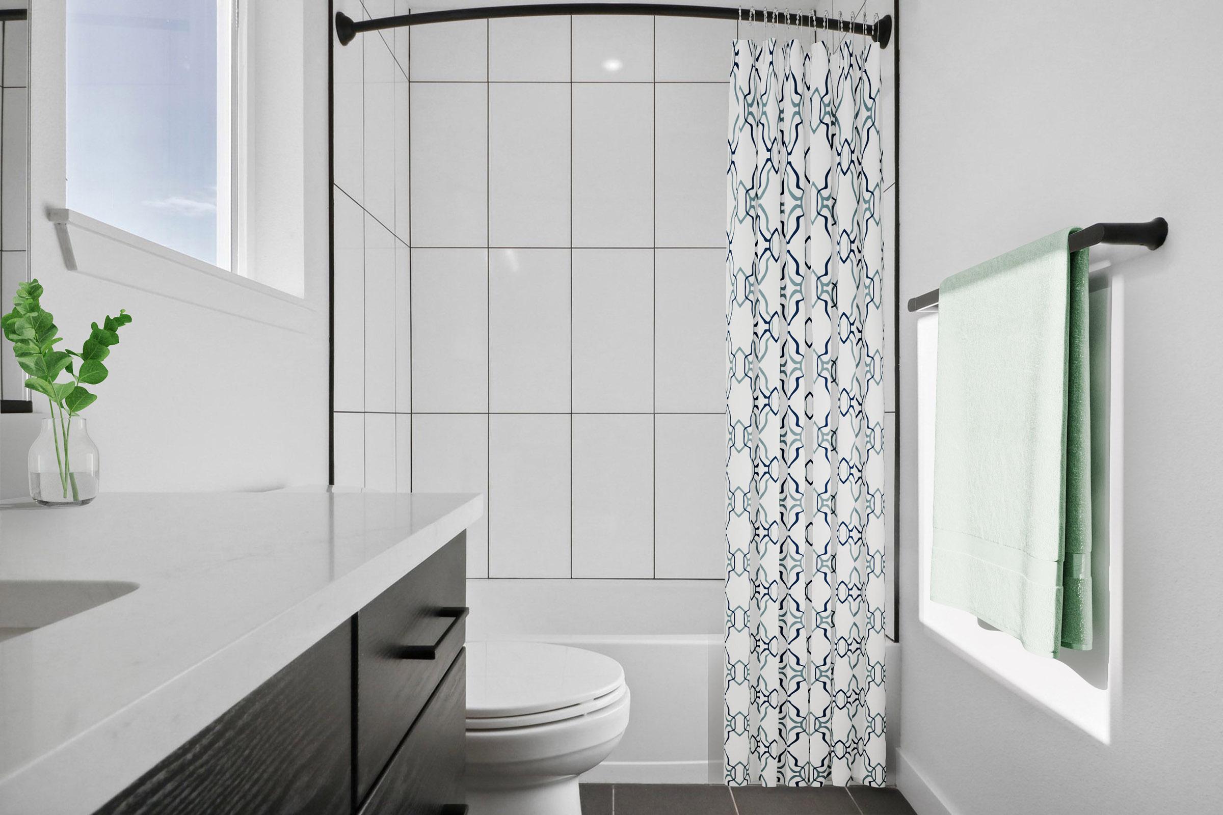 A modern bathroom featuring a white tiled shower with a patterned blue and white curtain, a white toilet, and a black vanity with a marble countertop. A green towel hangs on a rod, and a small plant is in a vase next to the sink. Natural light flows in from a window.