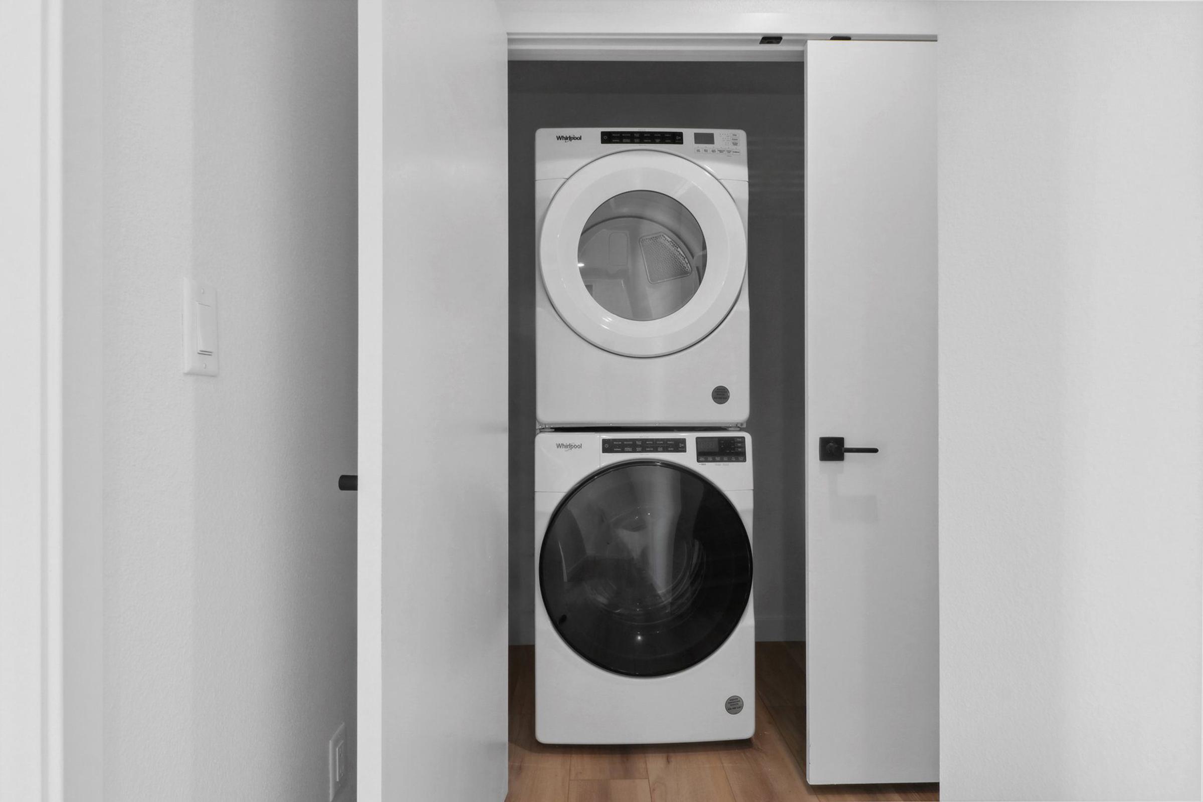 A modern laundry area featuring a stacked washer and dryer in a sleek white finish, situated against a light-colored wall. The appliances are framed by a doorway, creating a tidy and organized appearance. The floor is wood, adding warmth to the minimalist space.