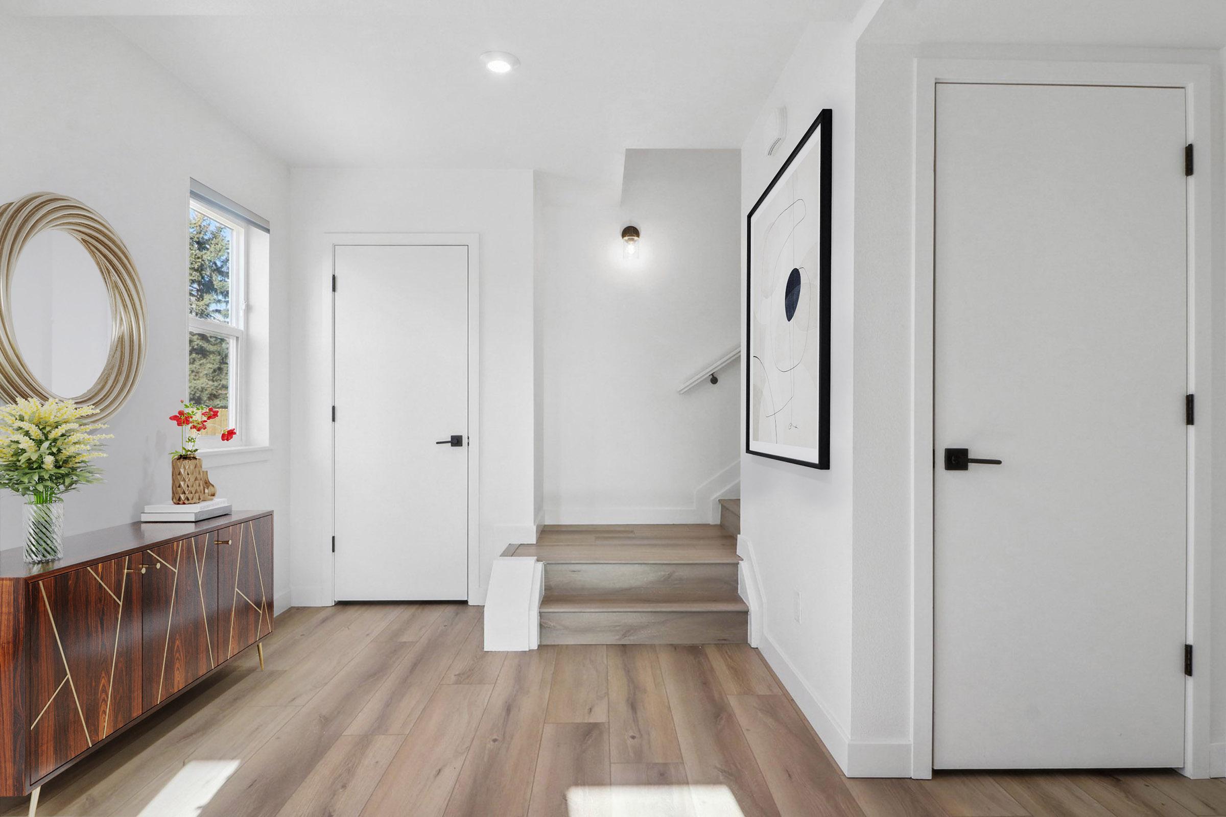 A bright and modern entryway featuring a small staircase leading upwards, a round mirror on the wall, a decorative vase with flowers on a stylish sideboard, and a black-framed artwork. The walls are white, and there is a door on the right, adding to the clean and inviting atmosphere.