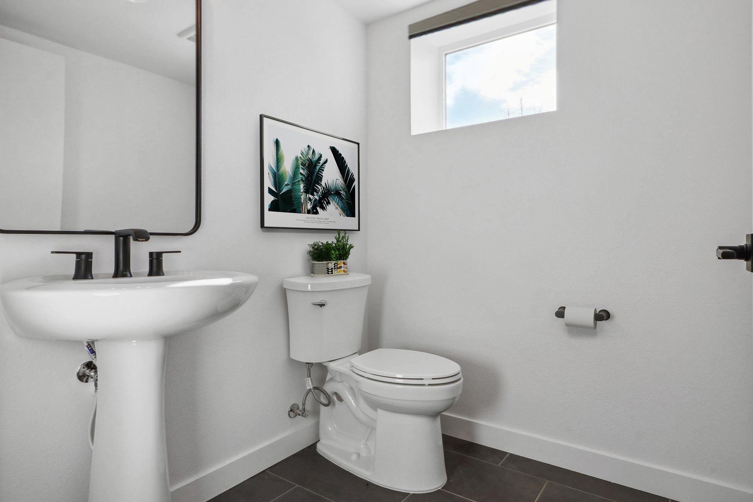 A modern bathroom featuring a white sink and toilet, a small window providing natural light, and a framed plant print on the wall. A potted plant sits on the toilet tank, and there's a roll of toilet paper on the wall. The walls and tiles are neutral shades, creating a clean and minimalist look.