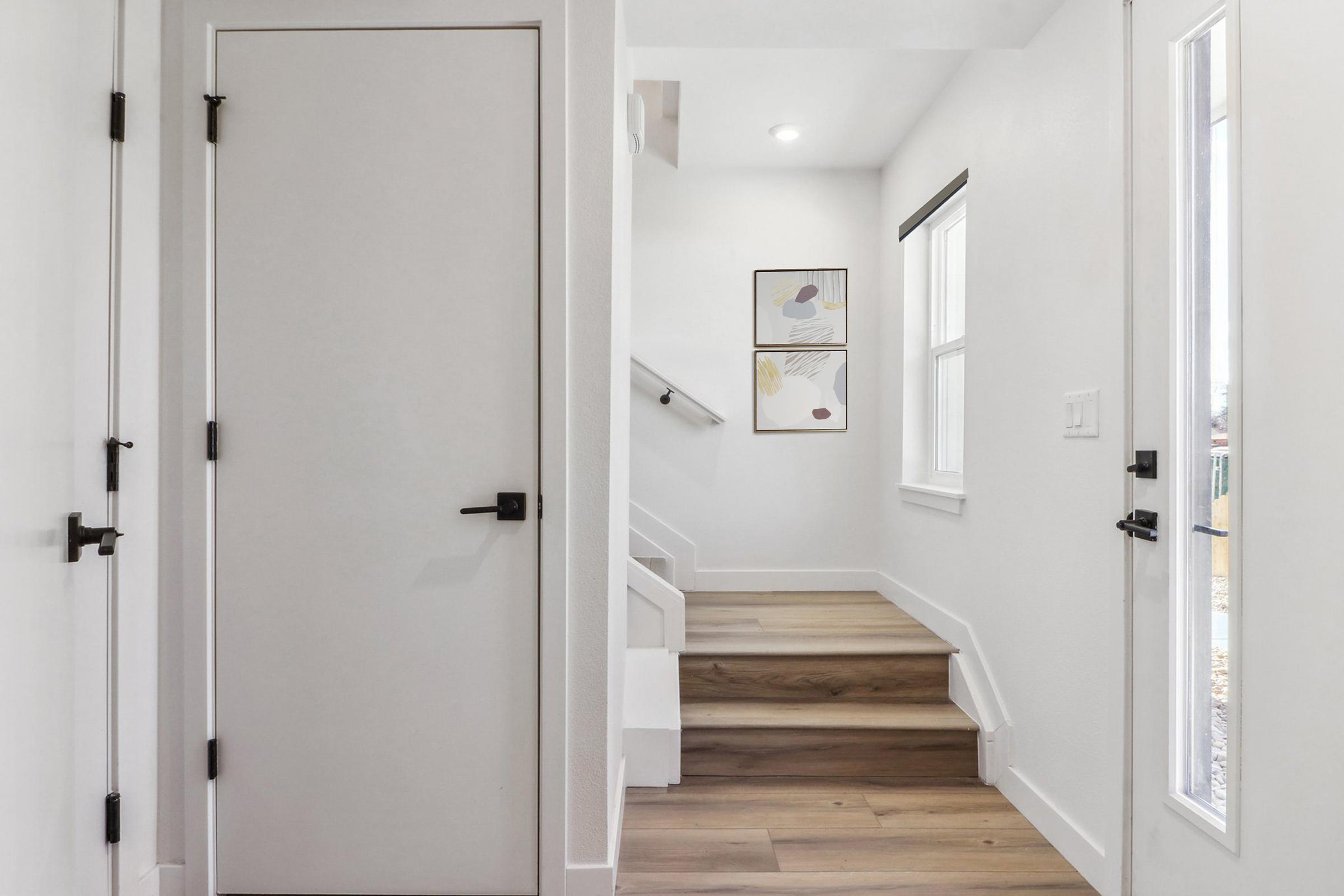 A well-lit hallway featuring light-colored walls, wooden flooring, and a closed door on the left. A set of stairs leads upward, with natural light coming through a window on the right. Two framed artworks are positioned on the wall across from the stairs.