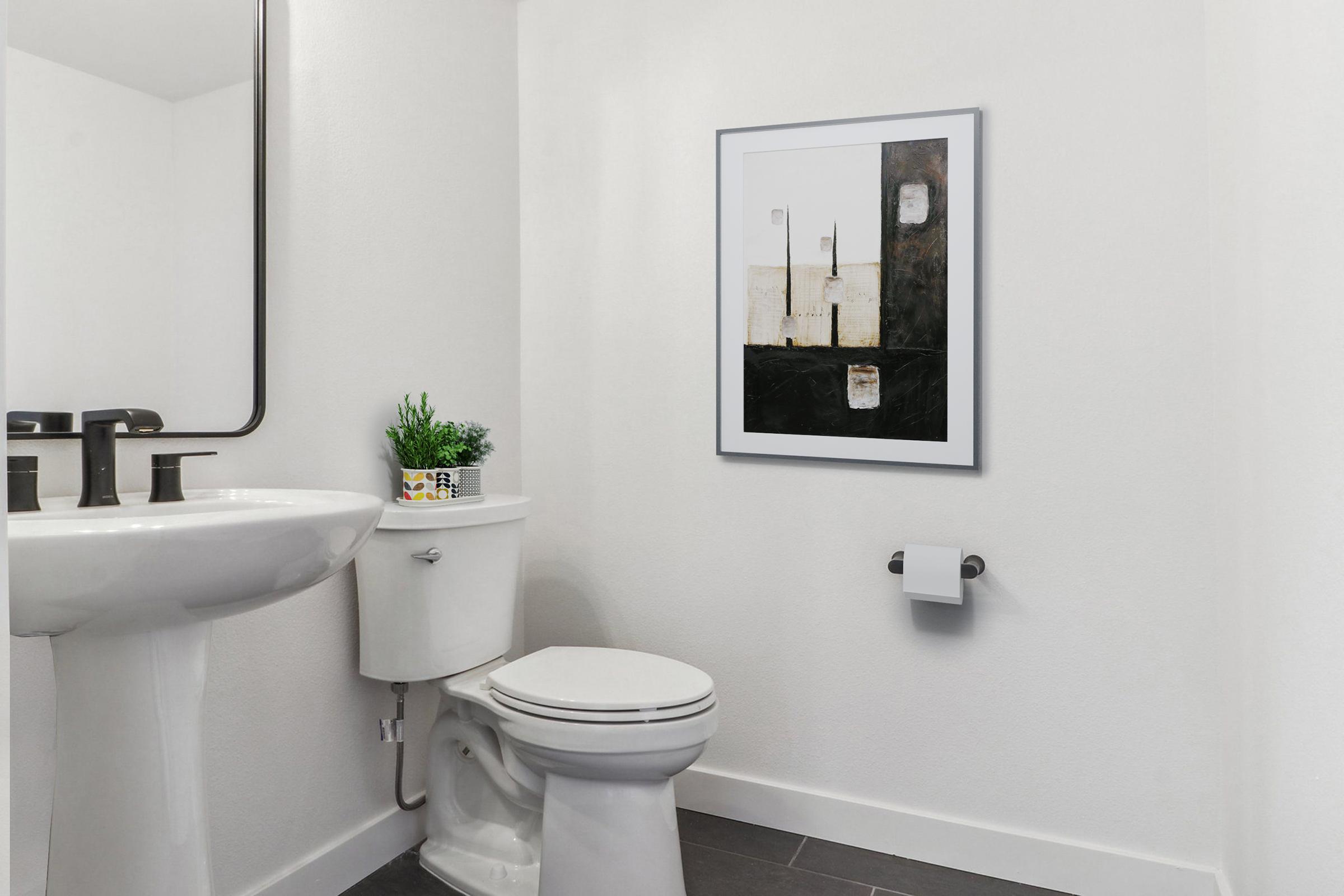 A modern bathroom featuring a white sink and a toilet, with a large mirror above the sink. A framed abstract artwork hangs on the wall next to the toilet, complemented by a small plant on the sink. The walls are painted white and the floor has dark tiles, creating a clean, minimalist aesthetic.