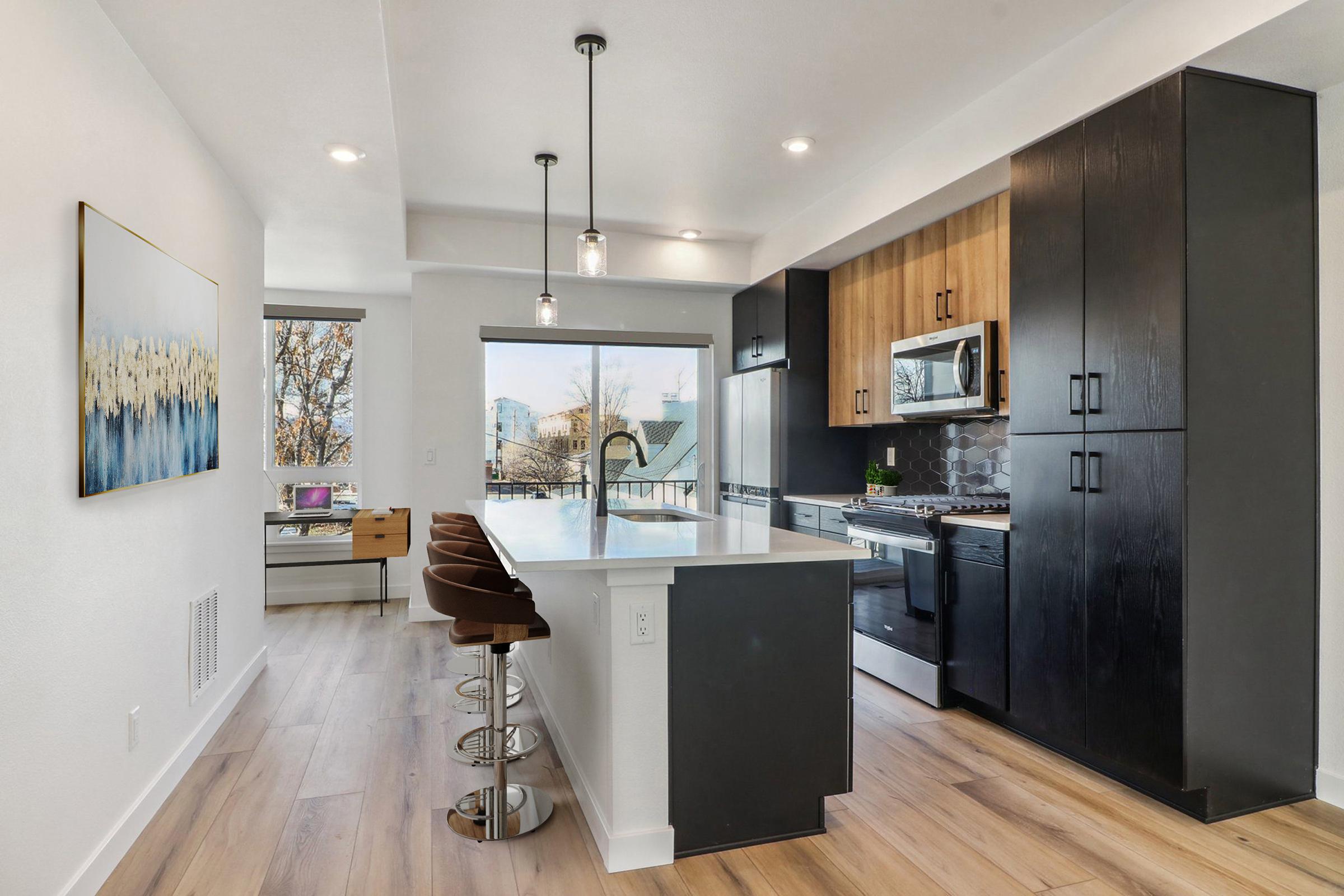 Modern kitchen featuring a white island with stools, sleek cabinetry in dark wood and light finishes, stainless steel appliances, and large windows providing abundant natural light. A small workspace is visible in the background, showcasing a minimalist design aesthetic.