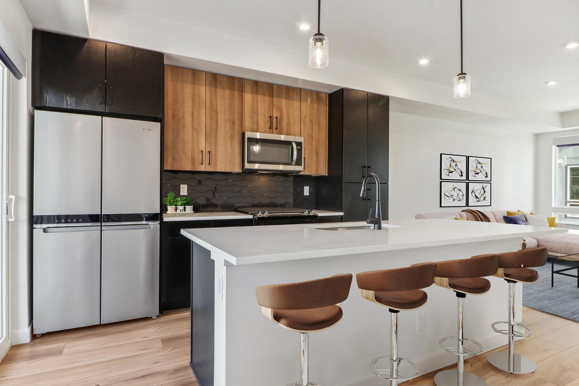 Modern kitchen featuring a sleek design with a combination of dark cabinets and light wood accents. Stainless steel appliances, including a refrigerator and microwave, are visible. A white island with three stylish bar stools is in the foreground, and a comfortable living area is seen in the background.