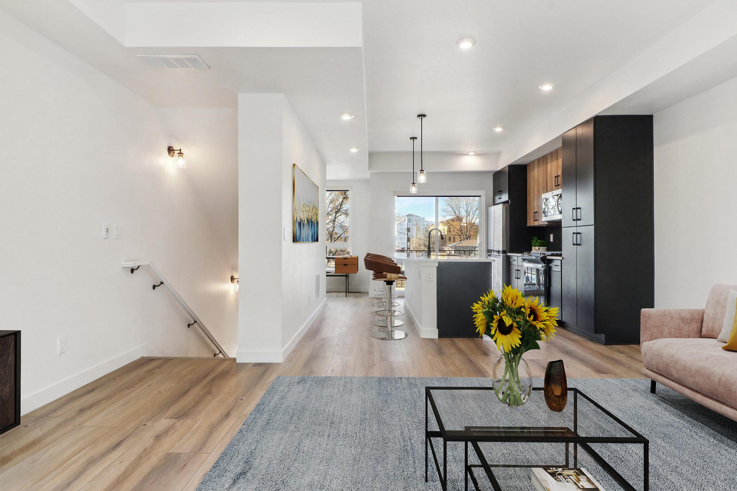 A modern interior of a bright and spacious living area featuring a comfortable pink sofa, a glass coffee table with a vase of sunflowers, and an open kitchen with sleek cabinets. Large windows let in natural light, enhancing the contemporary design and warm wood flooring.