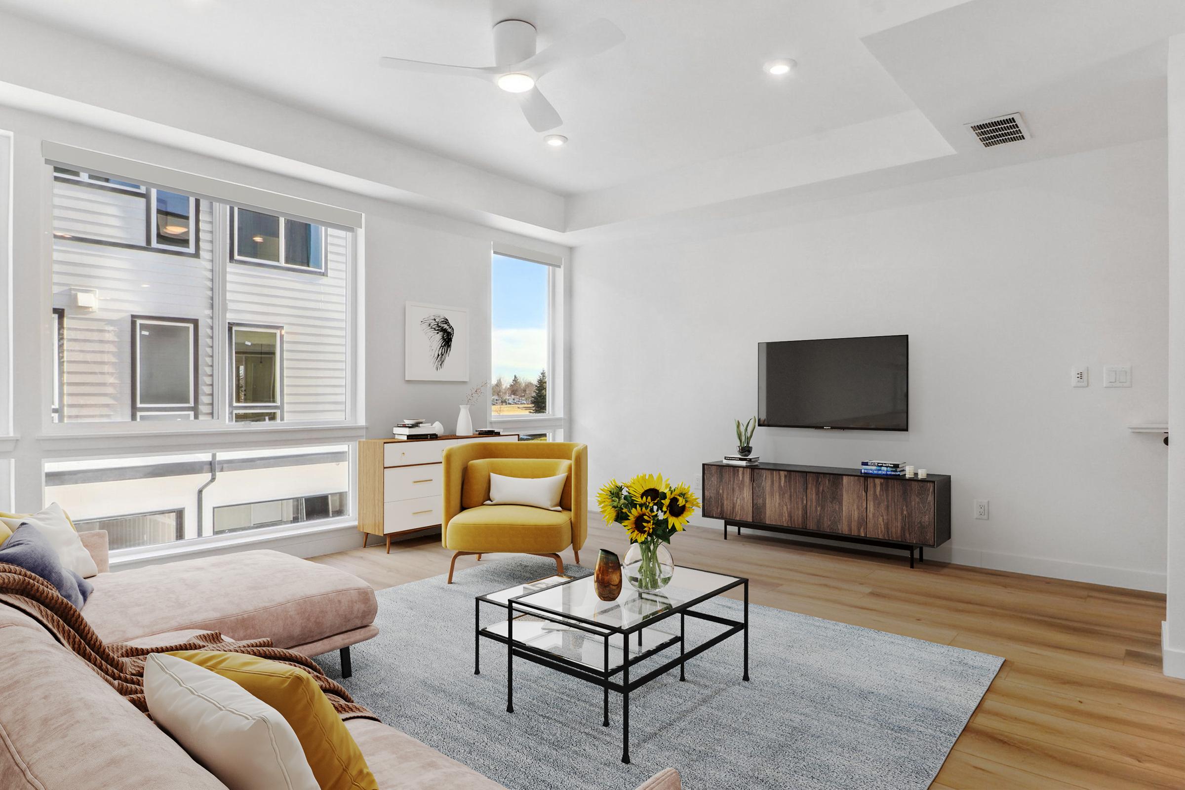 Bright and modern living room featuring a pink sofa, a yellow armchair, and a coffee table with a vase of sunflowers. Large windows allow natural light to fill the space, highlighting the neutral color palette and contemporary furnishings. A wall-mounted TV and sideboard complete the cozy atmosphere.