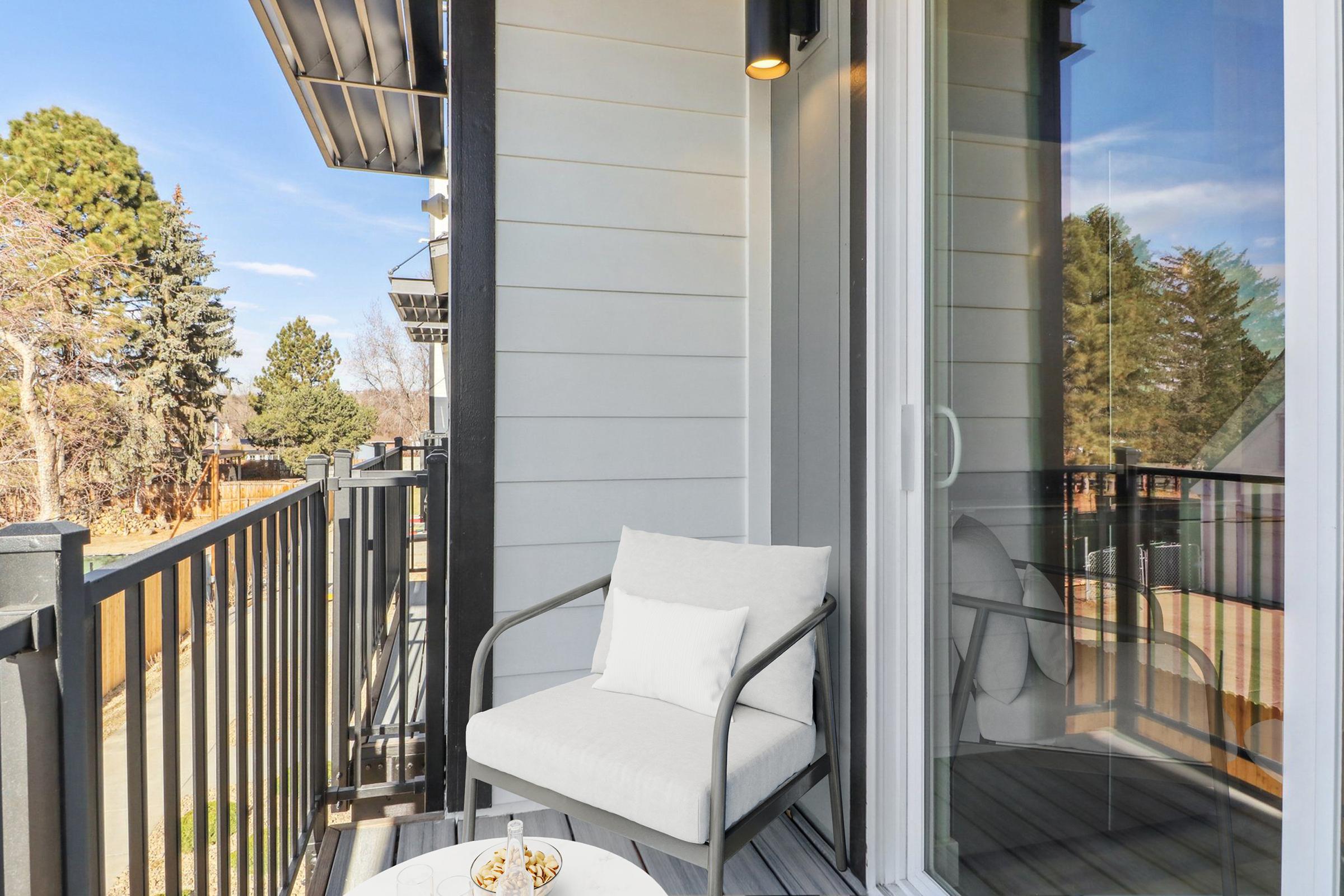 A cozy balcony with a light-colored chair and a small table. The setting includes greenery in the background and a clear blue sky, creating a peaceful outdoor space for relaxation. Sunlight adds warmth, enhancing the inviting atmosphere.