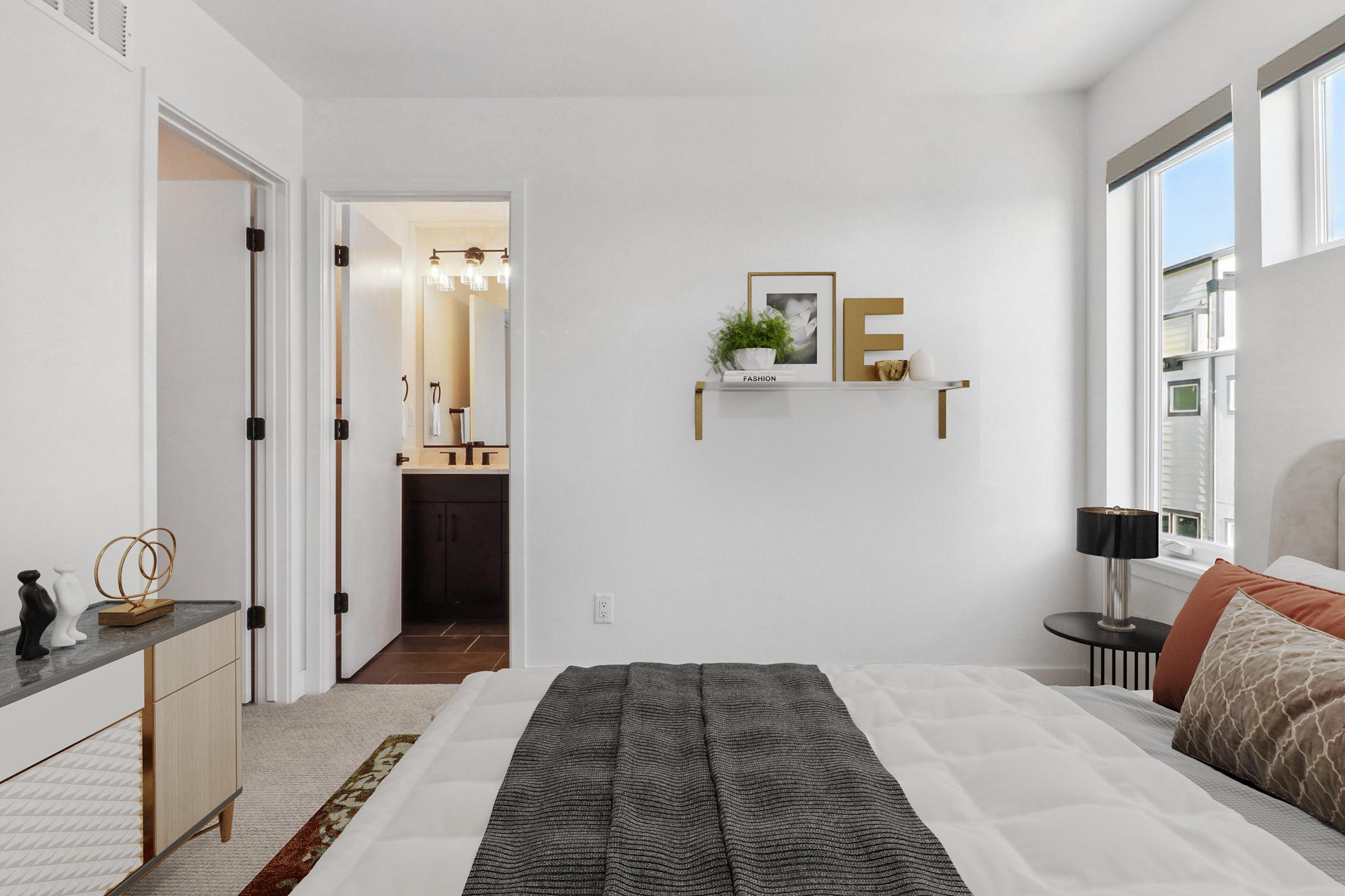 A modern bedroom featuring a neatly made bed with gray and orange pillows. A small shelf with decor items is mounted on the white wall. A doorway leads to a bathroom visible in the background. Natural light filters through large windows, creating a bright and spacious atmosphere.