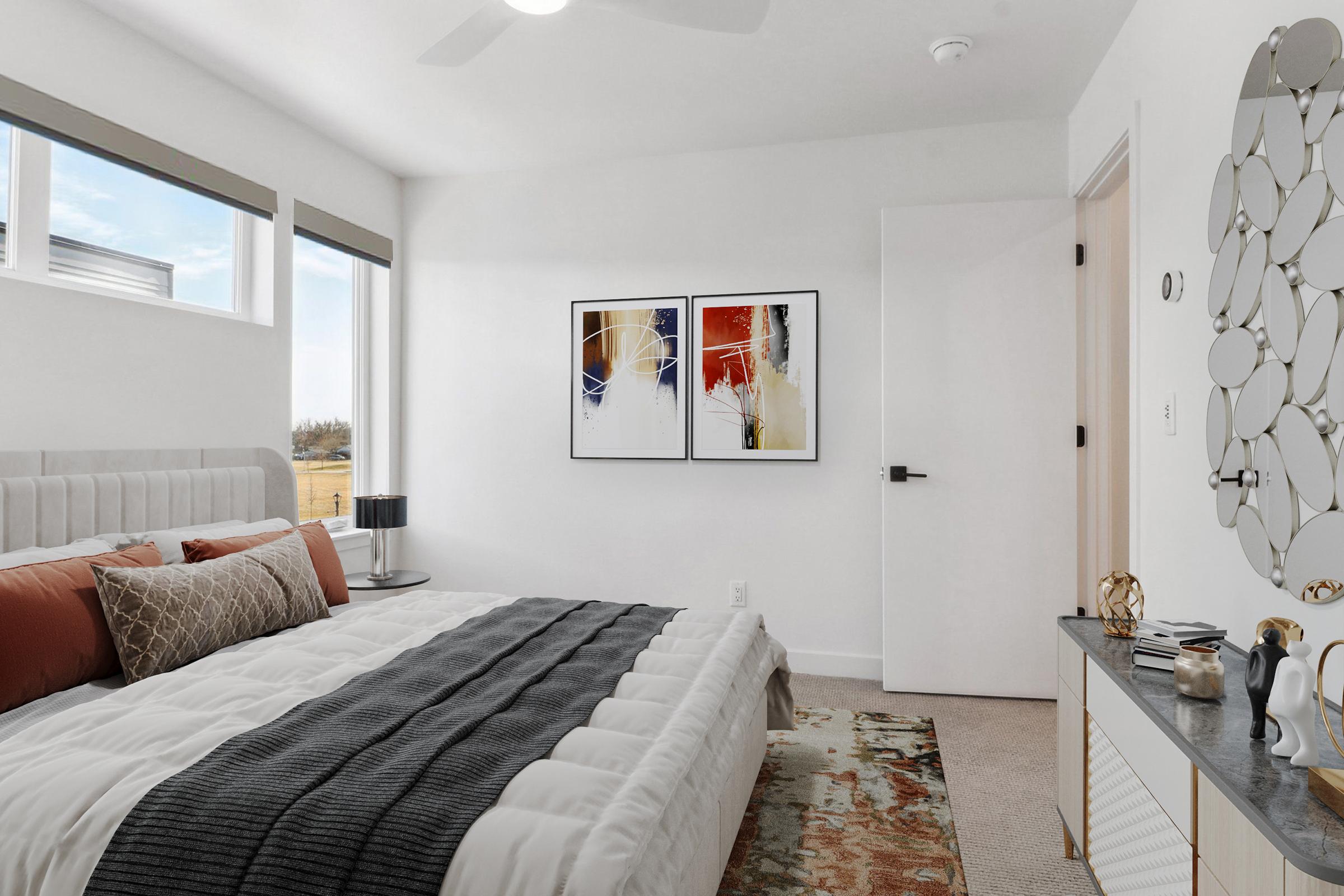 A modern bedroom featuring a large bed with a plush headboard, decorative pillows, and a textured gray throw. The room includes two abstract wall art pieces, a stylish nightstand with a lamp, a small table with decorative items, a circular mirror, and large windows allowing natural light to fill the space.