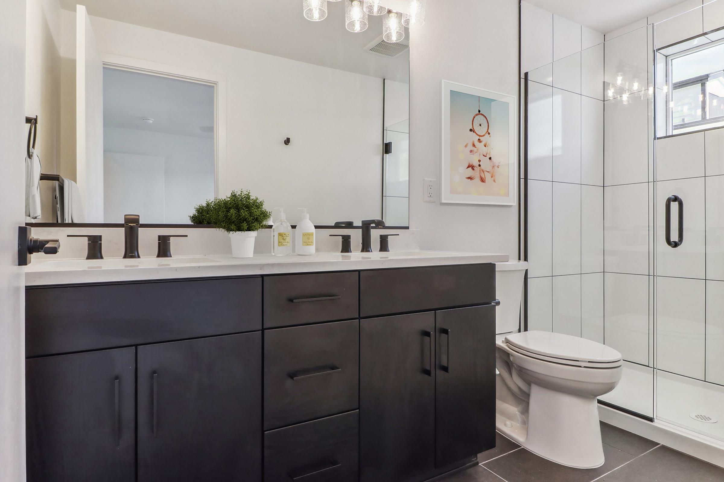 A modern bathroom featuring a sleek black double vanity with two sinks, stylish black fixtures, and a potted plant. There is a large mirror above the vanity, and a glass-enclosed shower nearby. The decor is minimalistic, with light-colored walls and subtle lighting fixtures.