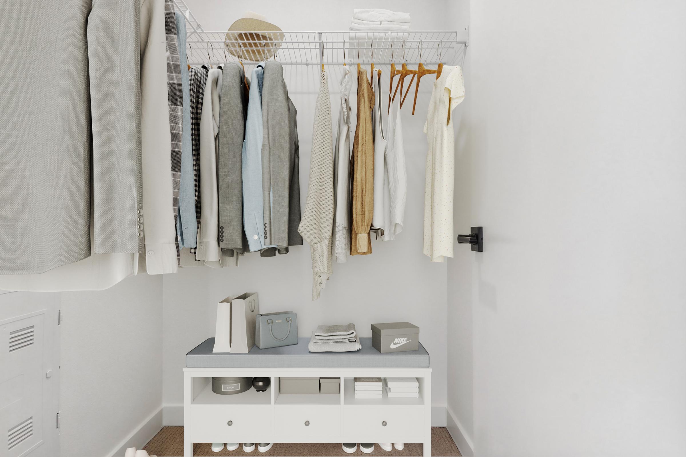 A neatly organized closet featuring hanging clothing items, including suits and casual wear. A white bench with storage boxes is at the bottom, and items like shoes and a handbag can be seen. The walls are a light color, and the overall space appears clean and modern, with a minimalist design.