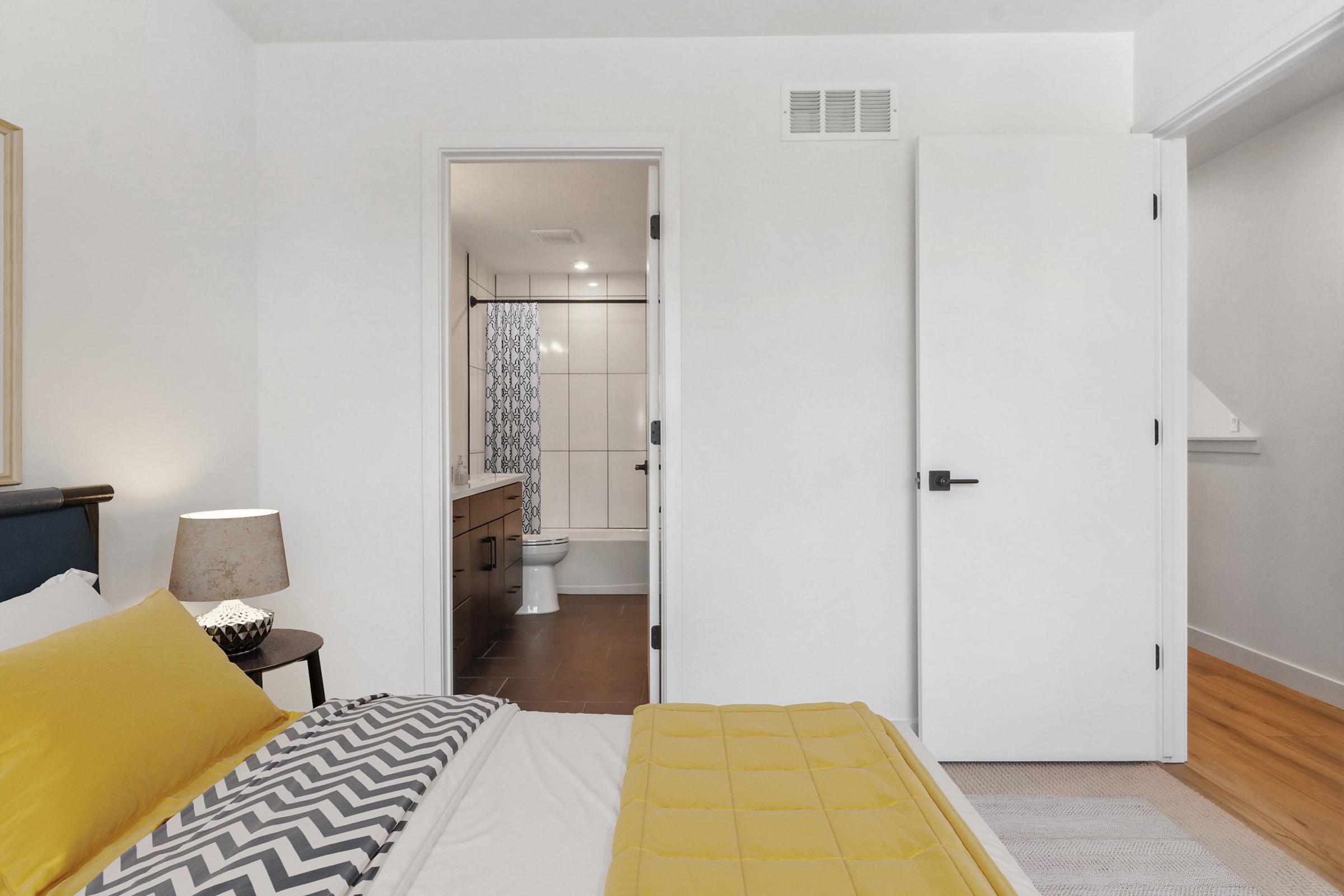 A cozy bedroom featuring a yellow and gray bedspread, a bedside lamp, and a view of an adjoining bathroom through an open door. The bathroom is modern with white tiles and a blue shower curtain. The room has light-colored wooden flooring and neutral-colored walls, creating a spacious and inviting atmosphere.