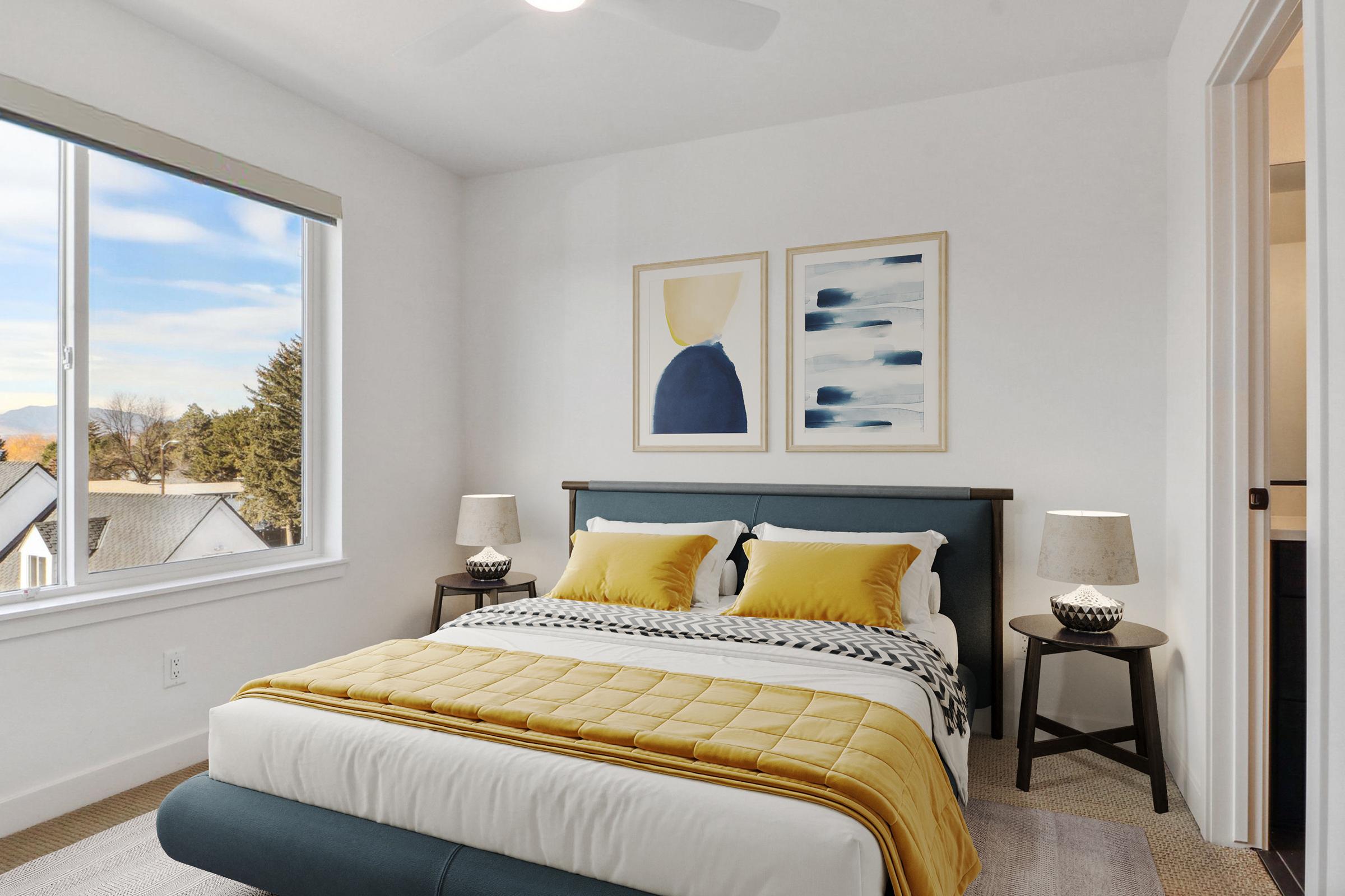 Cozy bedroom featuring a queen-sized bed with yellow and white bedding, accented by decorative pillows. Two framed art pieces hang above the bed. There's a window letting in natural light, and stylish bedside tables with lamps. Neutral walls and a light-colored rug complement the modern design.